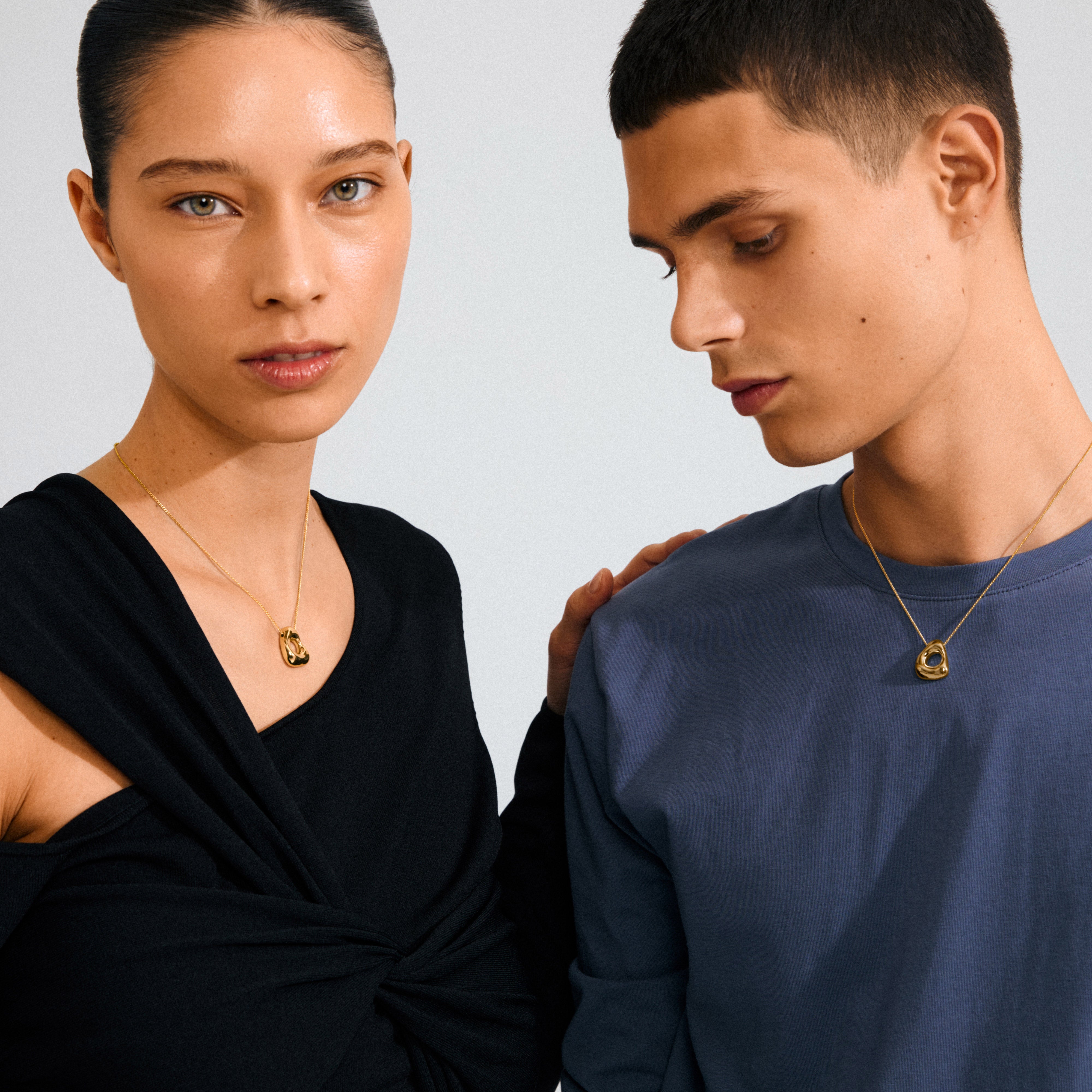 Two models wearing Pilgrim's FREE gold-plated necklaces; the woman's draped top contrasts with the man's muted shirt, highlighting the delicate, nature-inspired pendants.