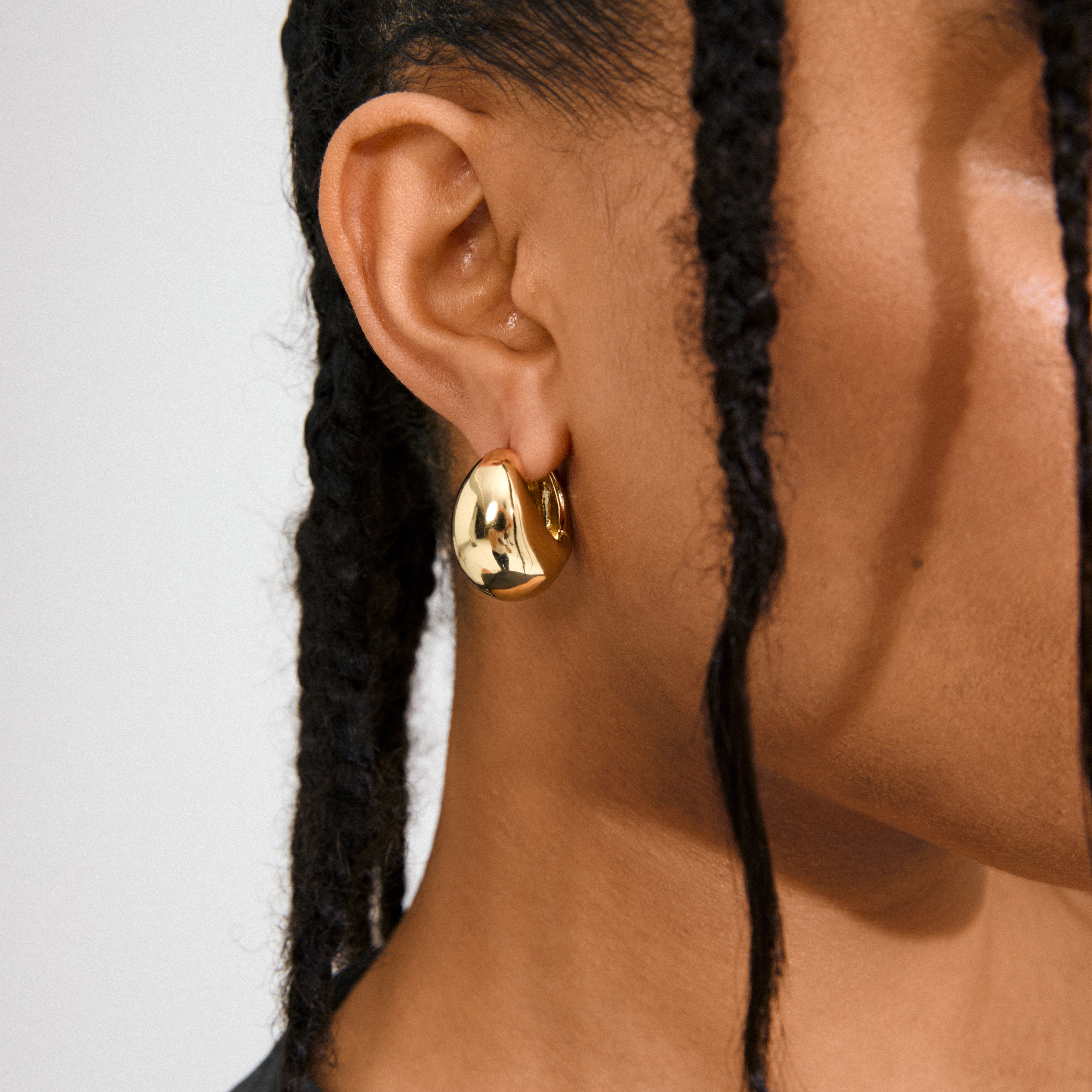 Close-up of a medium-dark skin ear wearing Pilgrim's FREE hoops gold-plated, featuring a chunky, uneven sculptural design, framed by thin, dark braids.