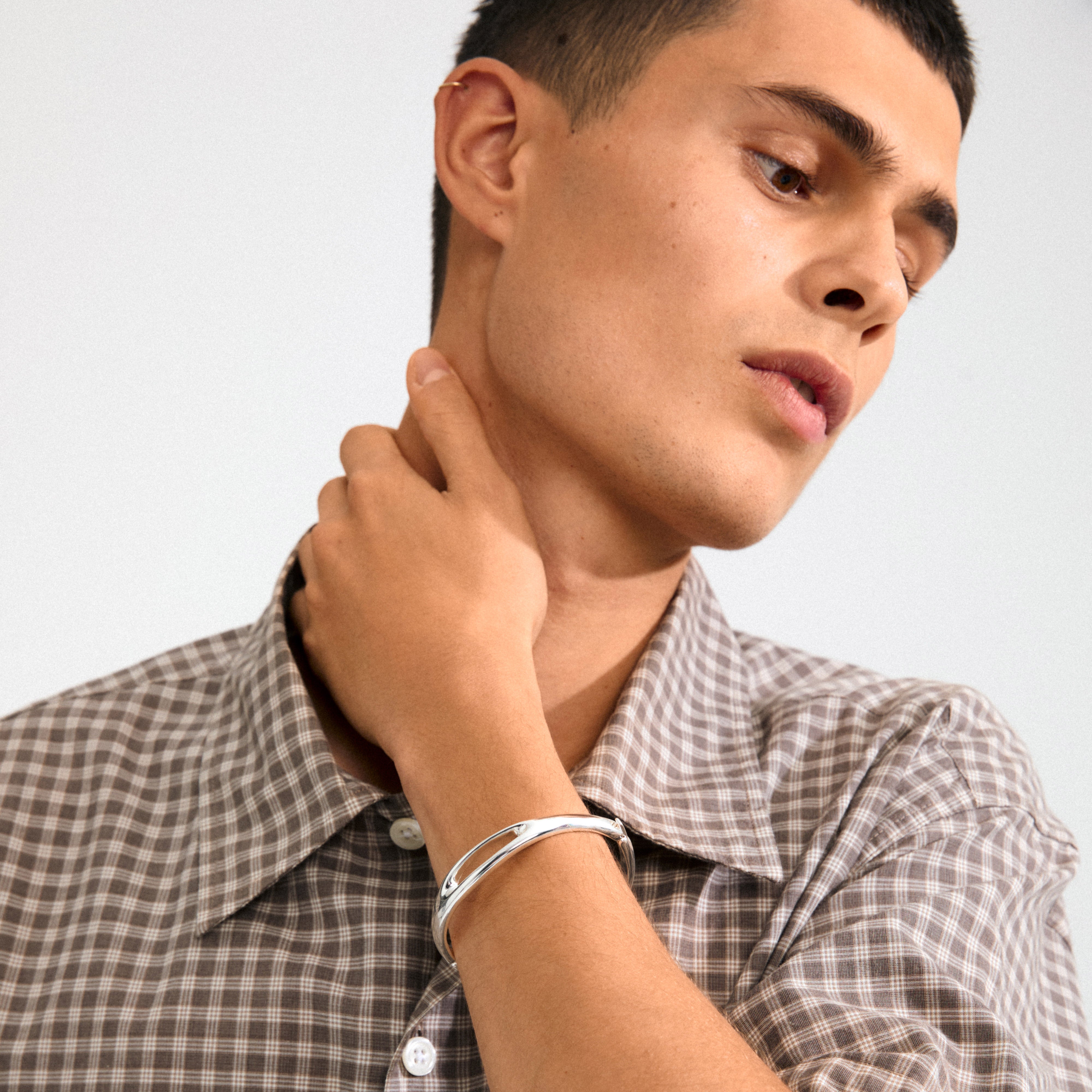 Young man wearing a gingham shirt and Pilgrim's FREE silver-plated bangle, showcasing its minimalist Scandi design and sculptural cut-out details on his wrist.
