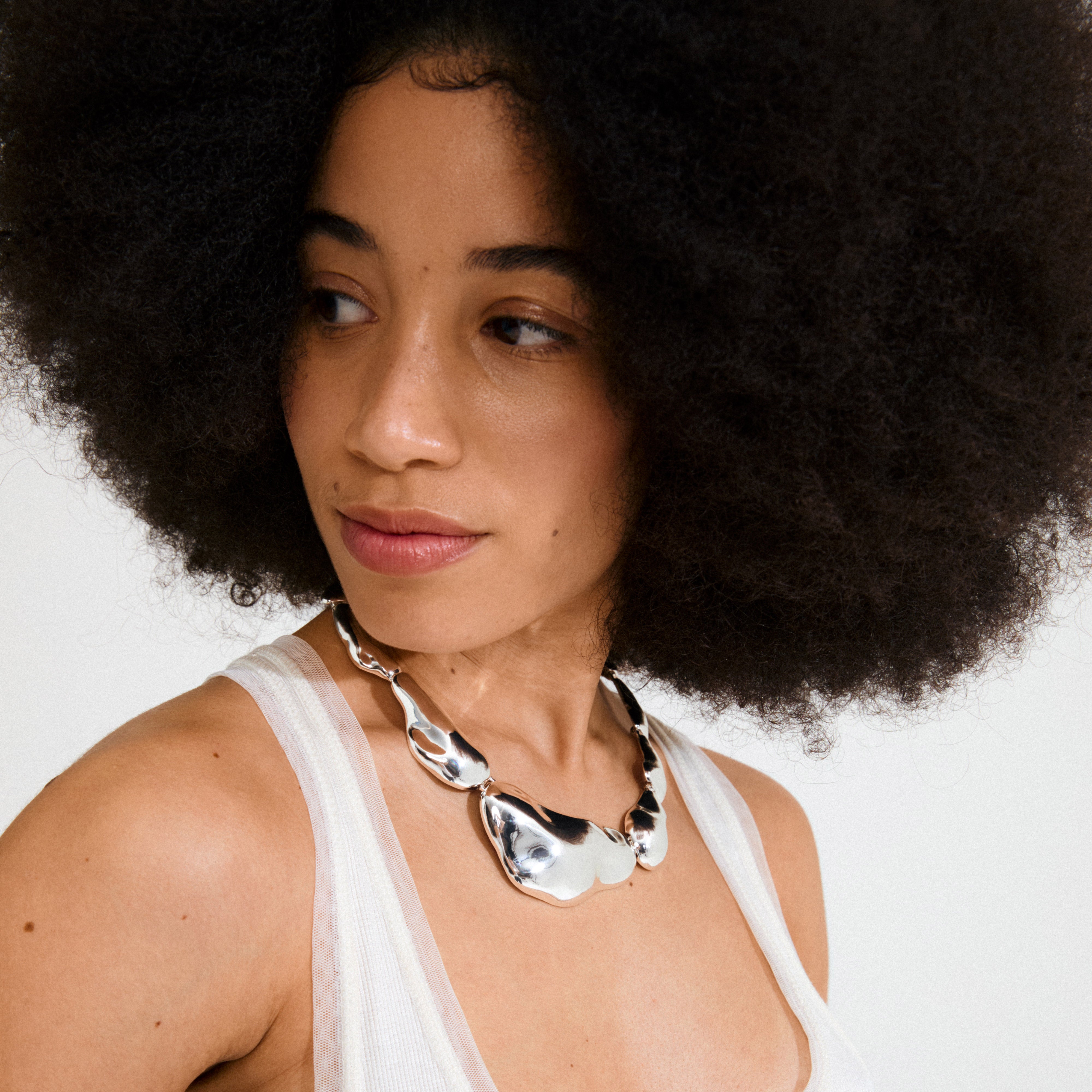 Young woman with a natural afro wears Pilgrim's FREE statement necklace, featuring chunky, silver-plated asymmetrical shapes, adding a bold, reflective charm to her look.