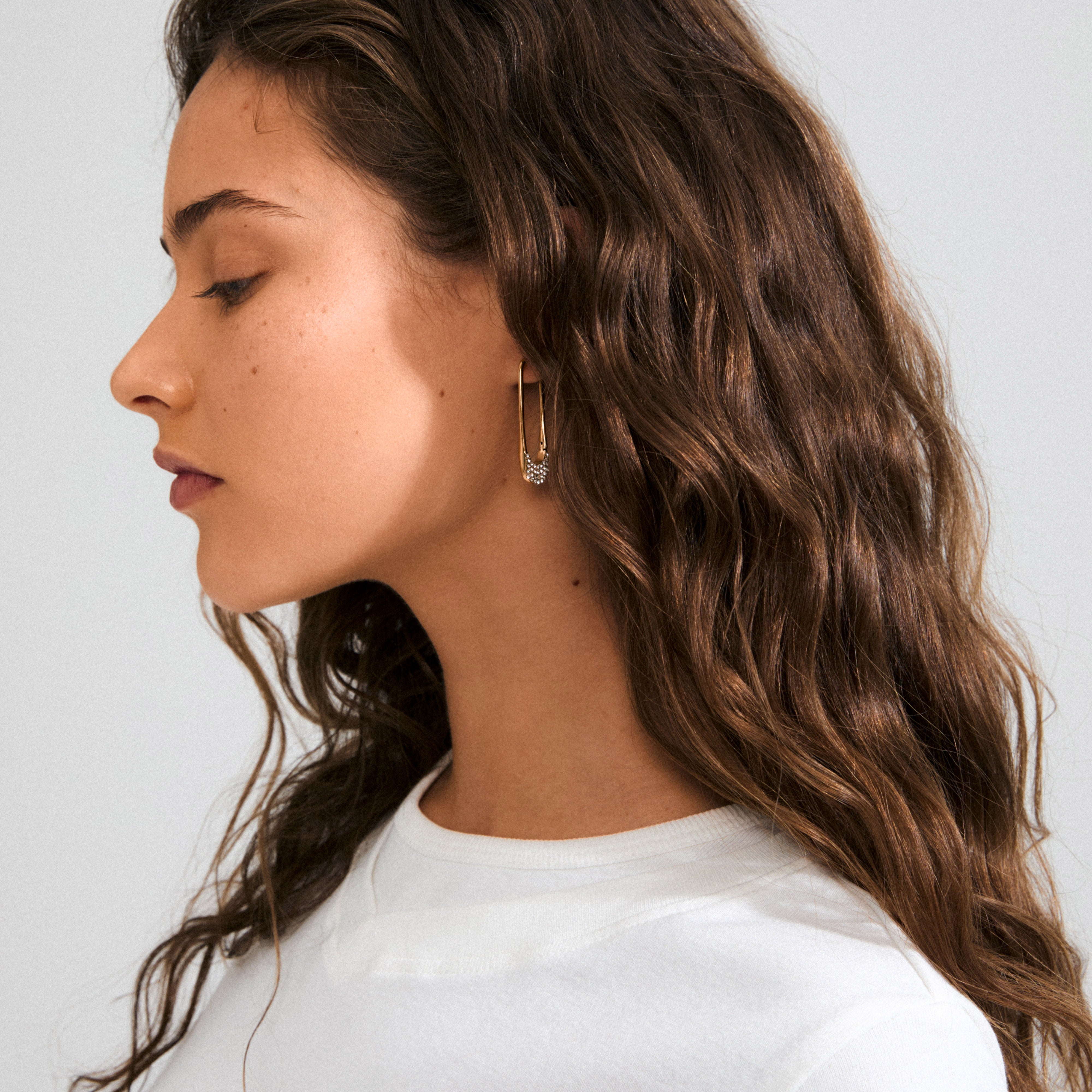 Side profile of a young woman wearing Pilgrim's INSTANT hoops, gold-plated safety-pin–style earrings with sparkling crystals, highlighting their edgy and glamorous design.