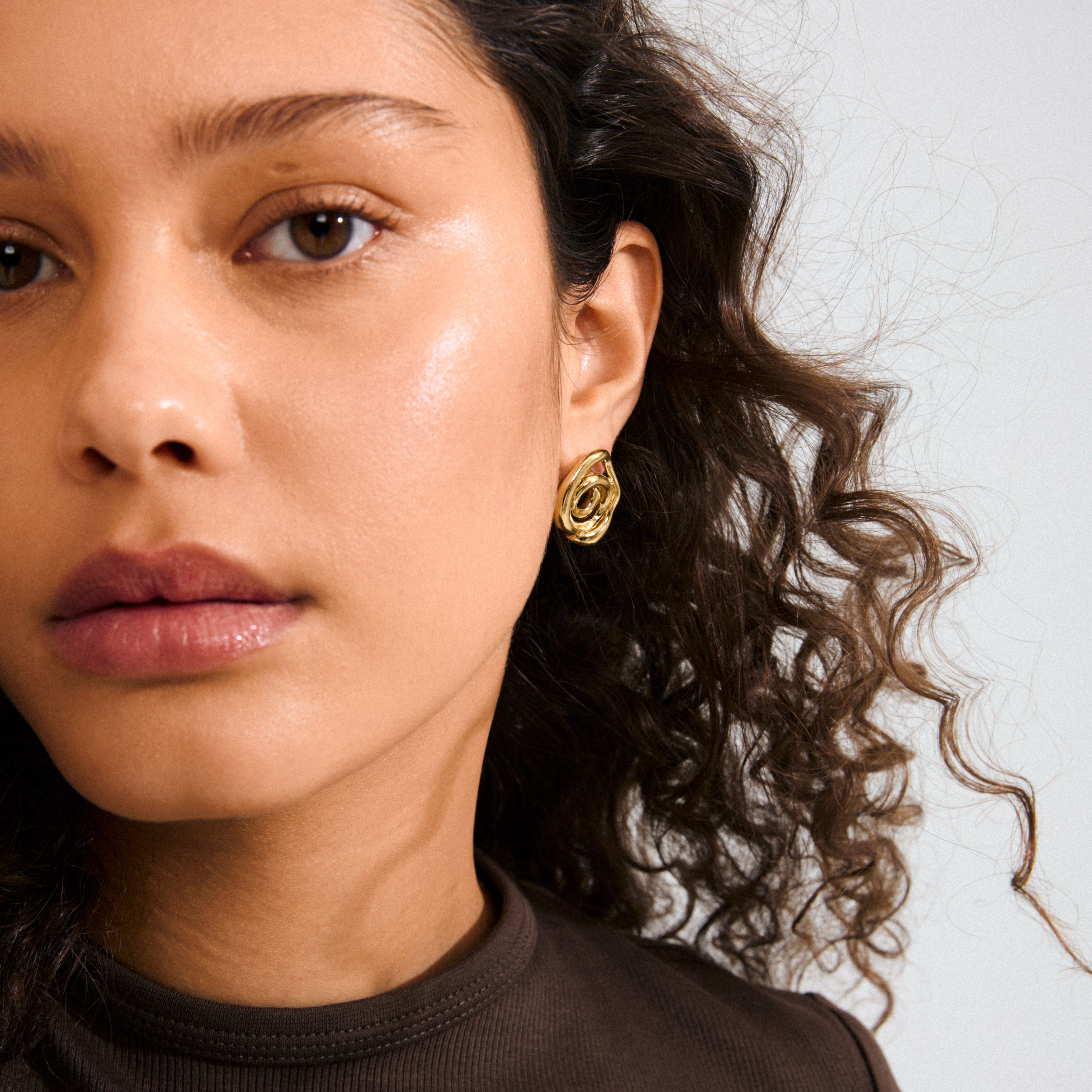 Close-up of a young woman wearing Pilgrim's UNFOLD gold-plated earrings, featuring a playful, sculptural swirl design that complements her dark curly hair and natural look.