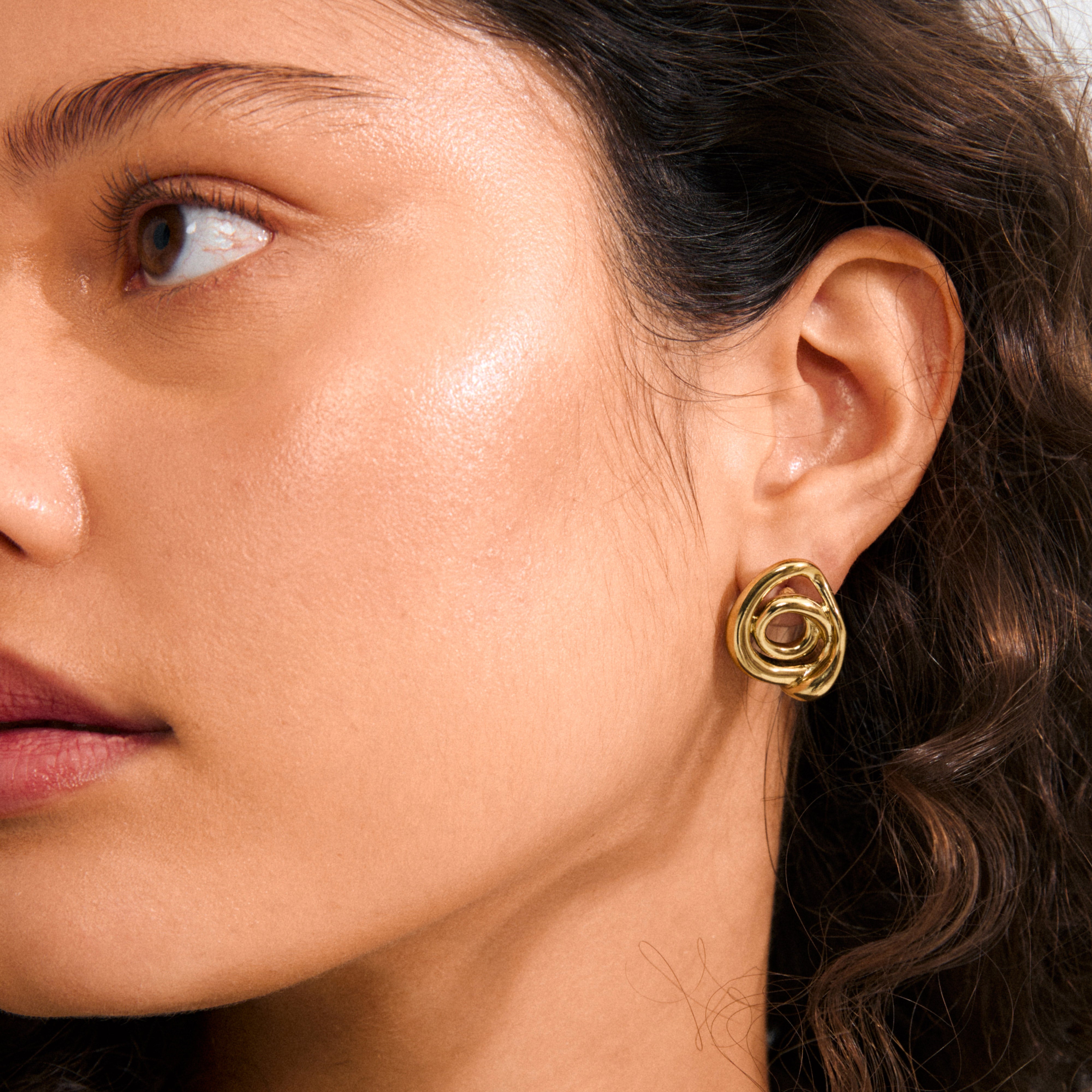Close-up of a person's ear wearing Pilgrim's gold-plated UNFOLD earring, featuring a sculptural, twisted knot design that adds an artistic, playful touch.