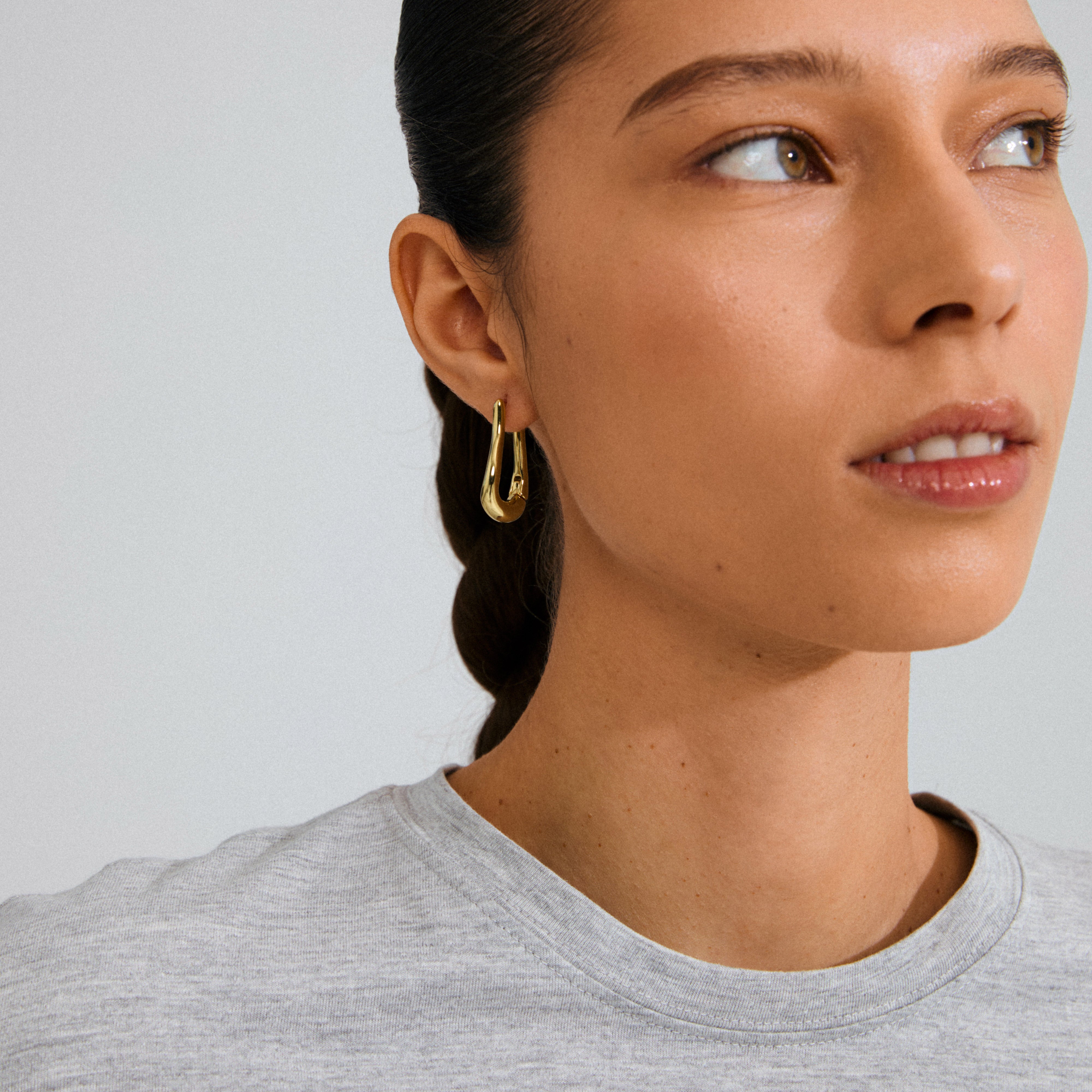 Woman wearing a light grey T-shirt and a single UNFOLD gold-plated hoop earring, showcasing its fluid, teardrop design and shiny finish.