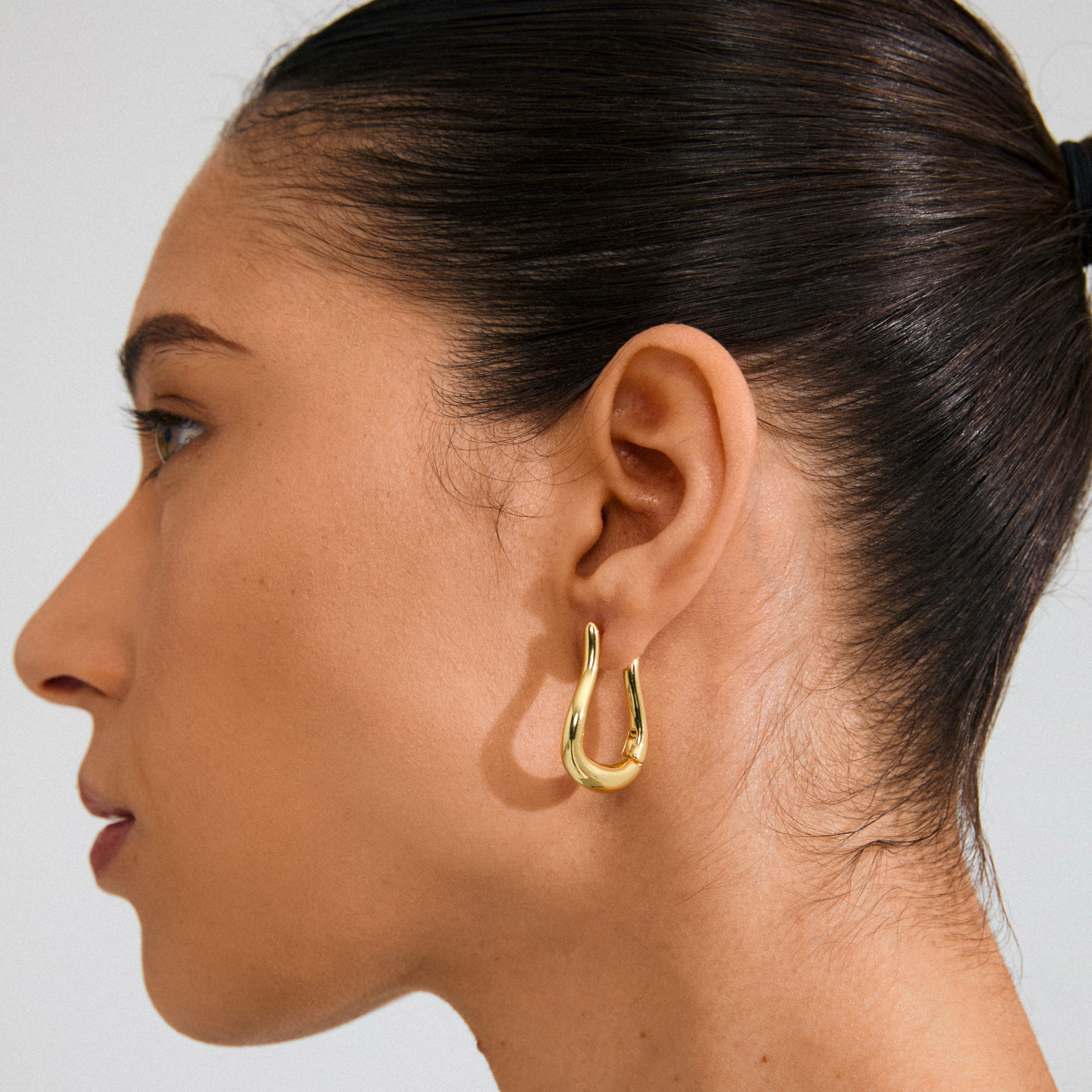 Side profile of a warm brown skin tone person wearing Pilgrim's UNFOLD gold-plated, teardrop-shaped hoop earring, showcasing its shiny, sculptural design.