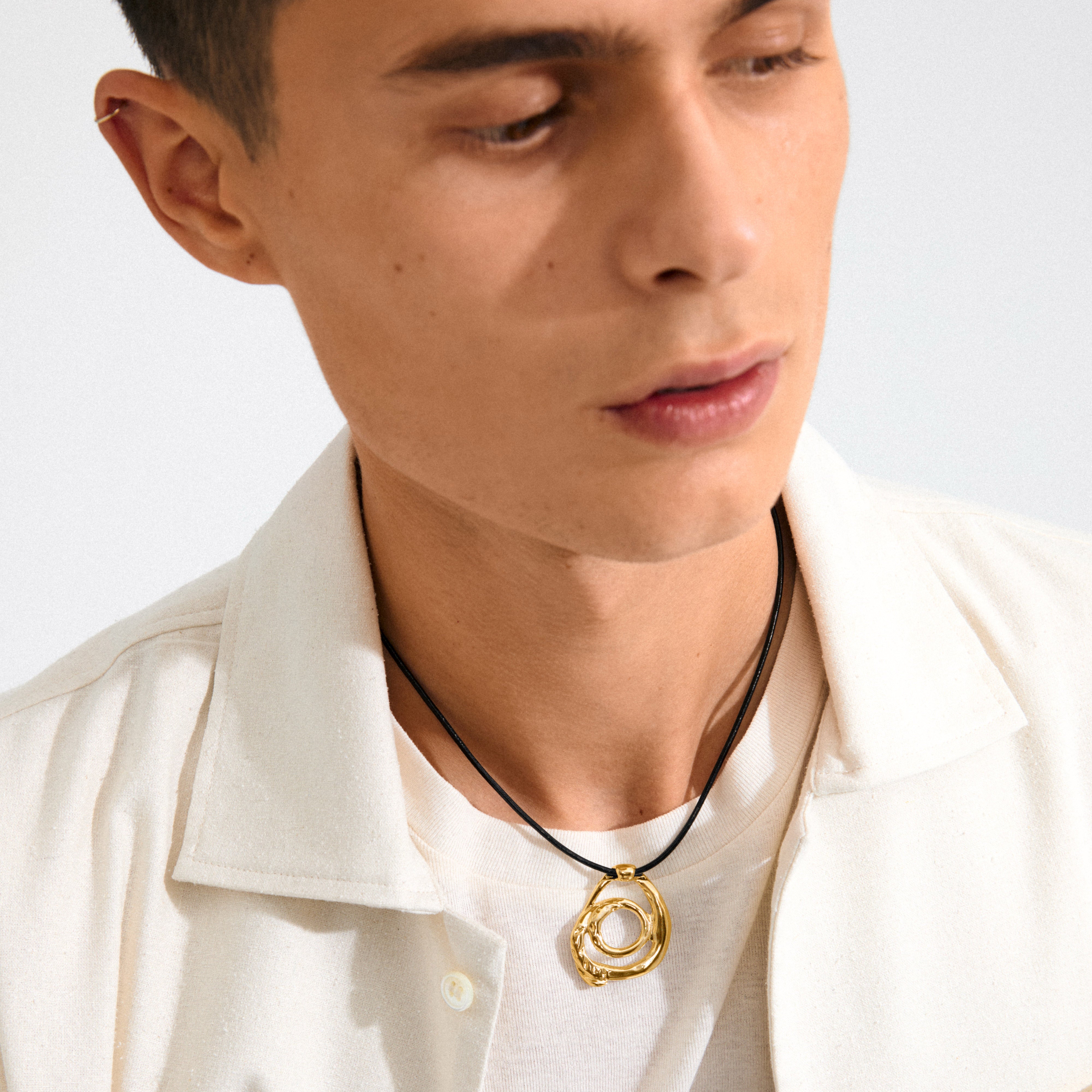 Young man wearing Pilgrim's UNFOLD necklace, featuring a gold-plated, twisted doodle pendant on a black leather cord, styled over a white tee and cream overshirt.