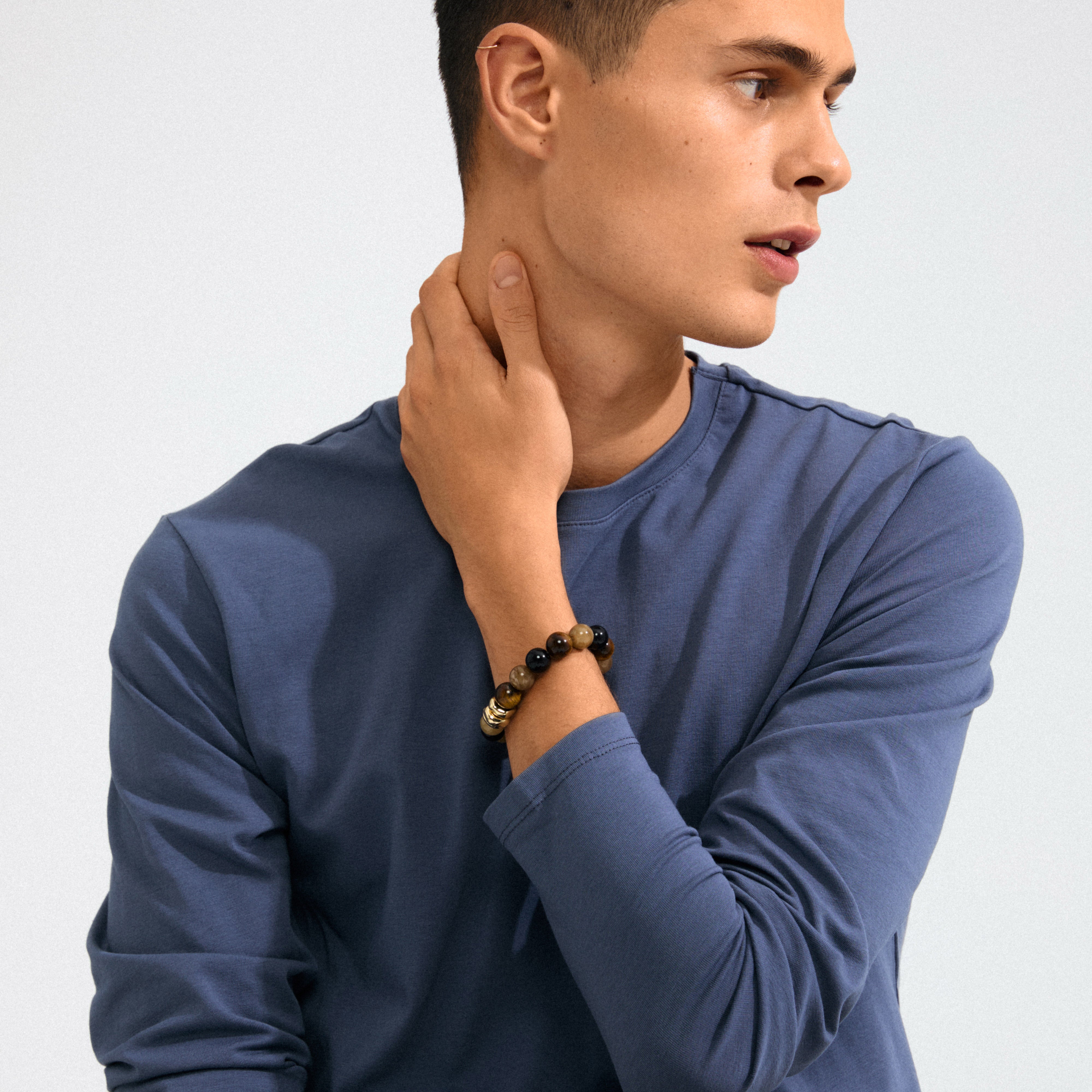 Young man wearing the UNFOLD bracelet, featuring chunky gold-plated, brown beads, and gold pendants, styled with a casual blue T-shirt.