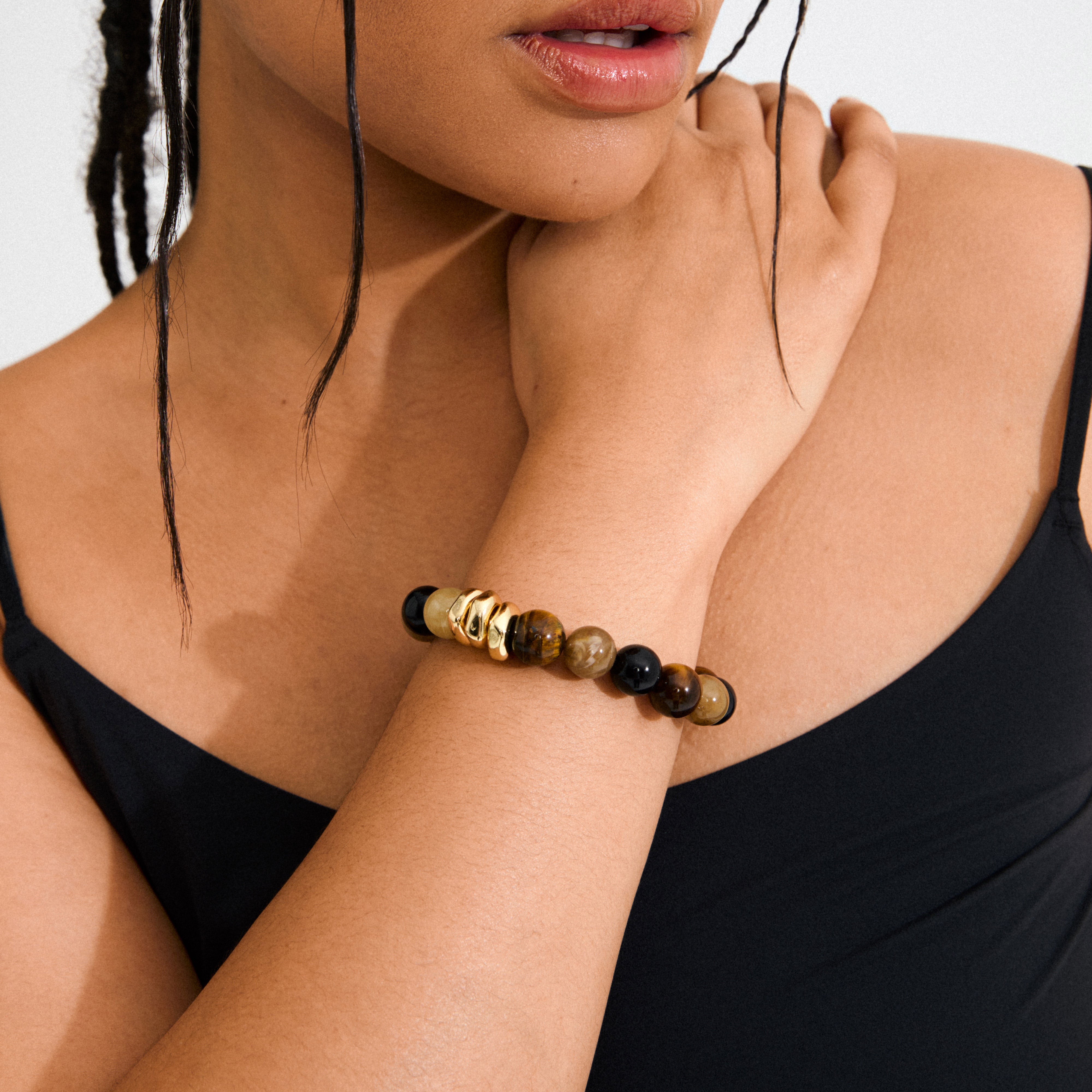 UNFOLD bracelet in gold-plated/brown, featuring chunky Tiger Eye beads and sculptural gold-tone accents on a raised wrist, complementing a black camisole.