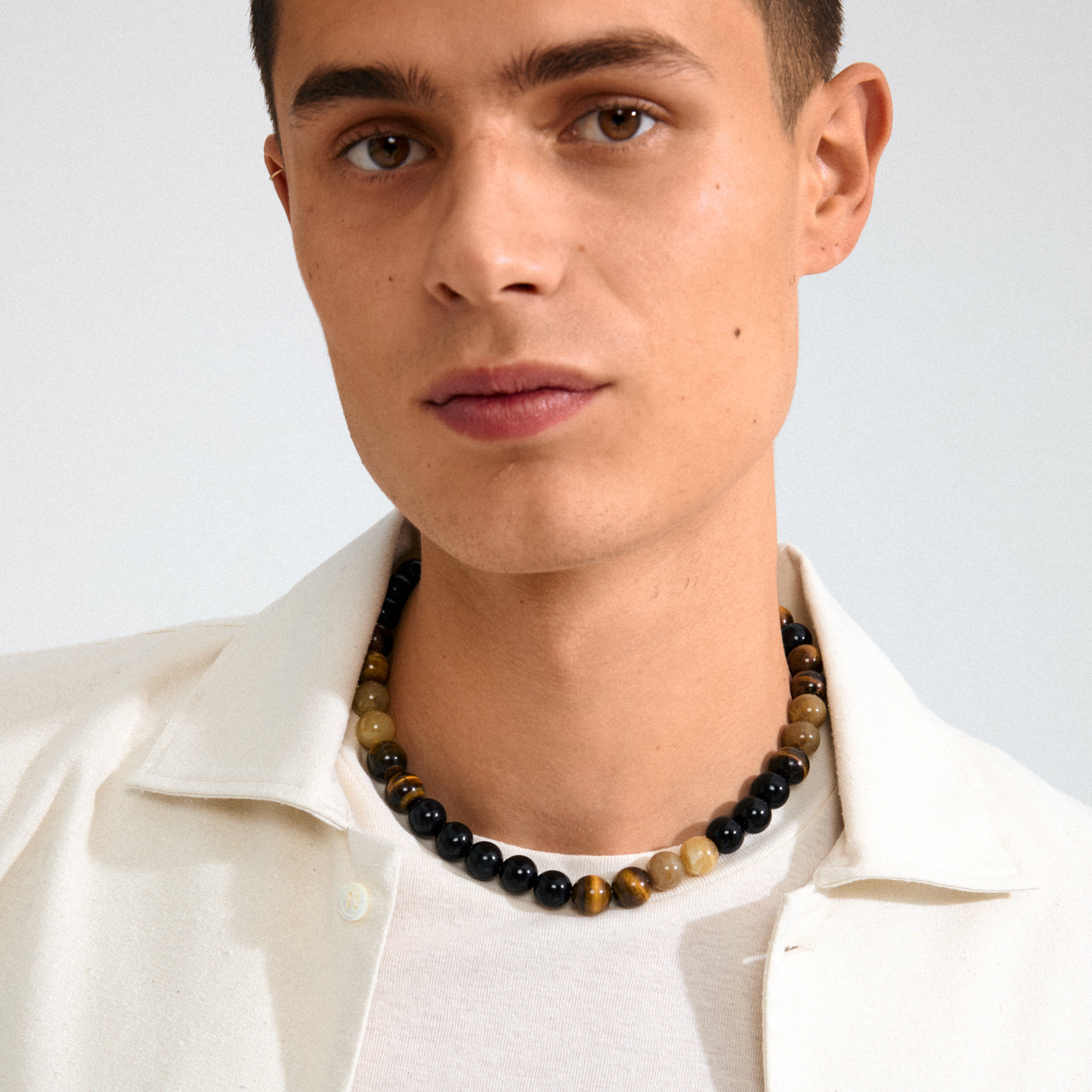 Young man wearing the UNFOLD necklace gold-plated/brown with chunky tiger-eye beads, adding an effortless, trendy touch to his autumn-inspired outfit.