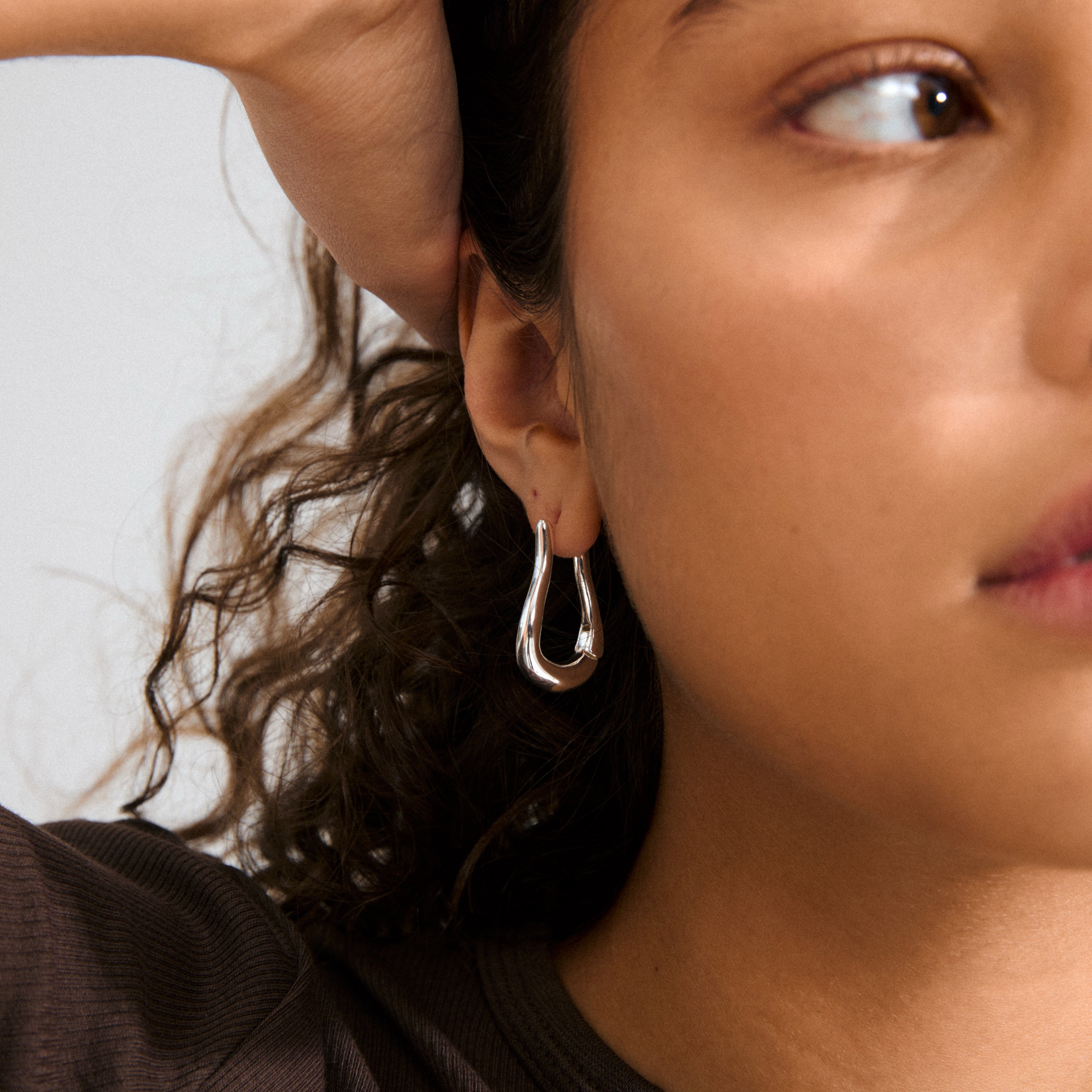 Silver-plated UNFOLD hoops on ear, showcasing their airy, teardrop shape and shiny finish, with hand holding back curly hair for stylish, urban elegance.