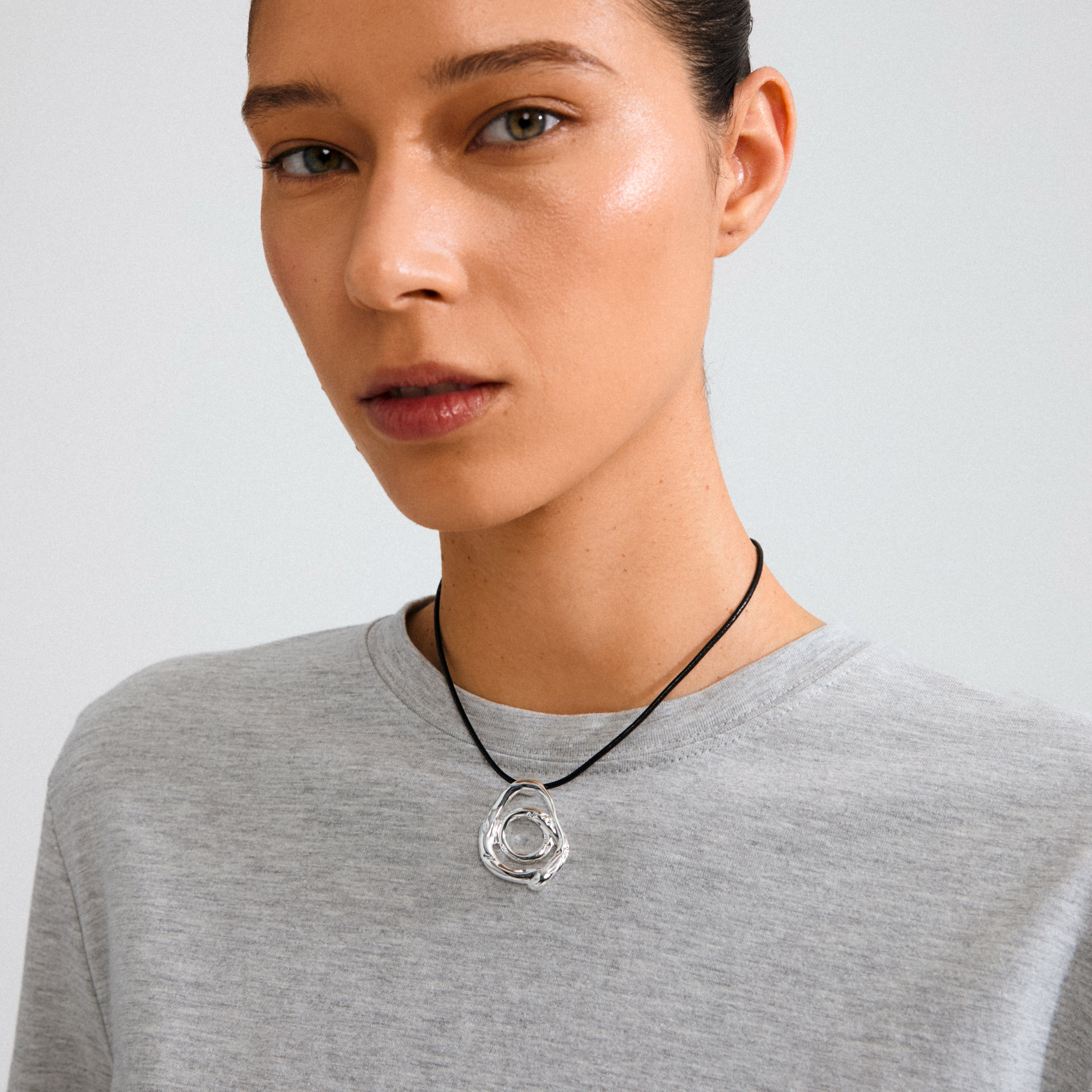 Young woman wearing the UNFOLD necklace, featuring a sculptural silver pendant on a black cord, styled over a light grey T-shirt for an artistic touch.