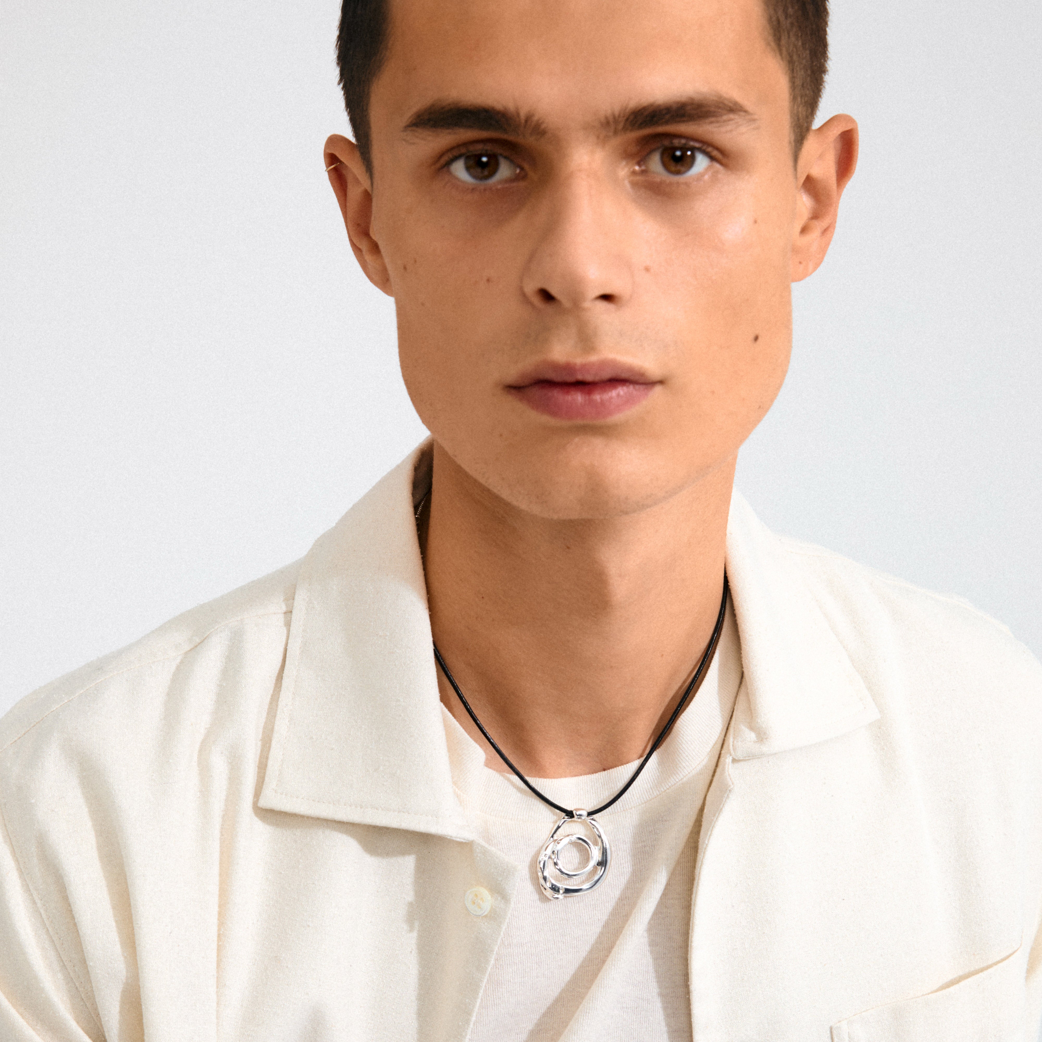 Young man wearing the UNFOLD necklace, a silver-plated piece with interlocking rings on a black cord, complementing his casual cream shirt and white T-shirt.