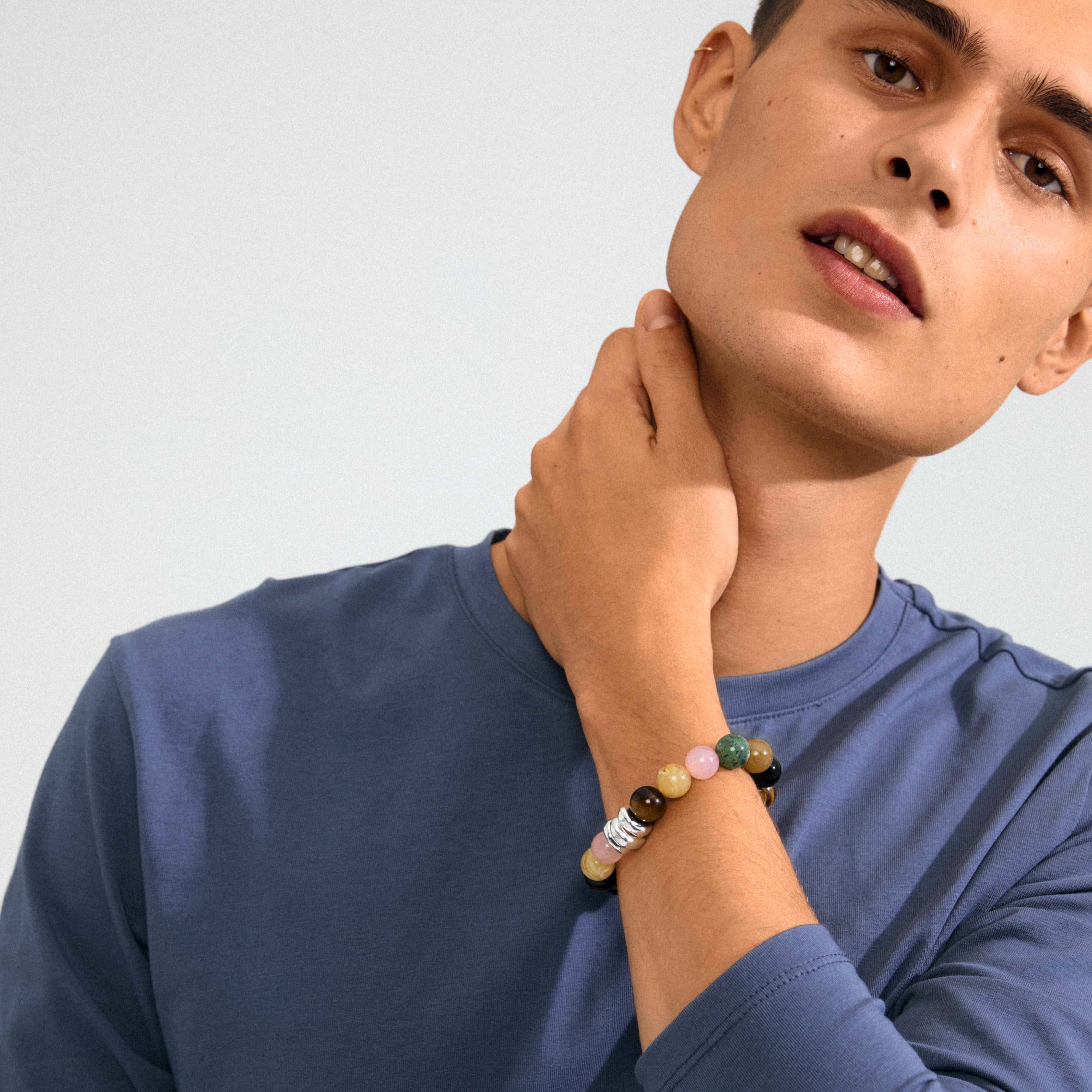 Young man wearing the UNFOLD bracelet by Pilgrim, featuring earthy-hued beads and silver-plated accents, complementing his muted blue shirt with a relaxed, confident pose.