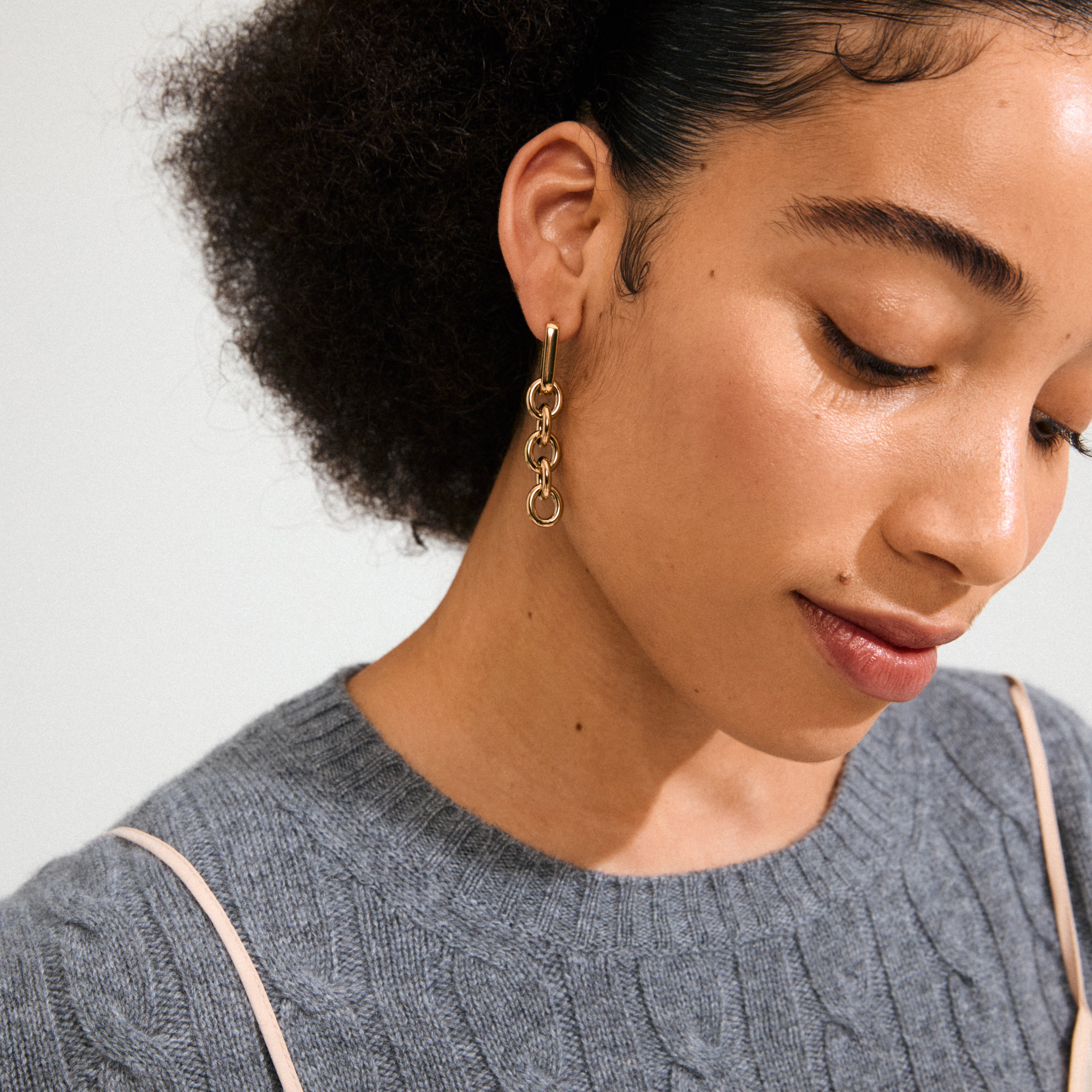 Woman wearing PHASE earrings gold-plated by Pilgrim, featuring interlocking rings in a cable chain style, complementing her grey cable-knit sweater and natural, textured hairstyle.