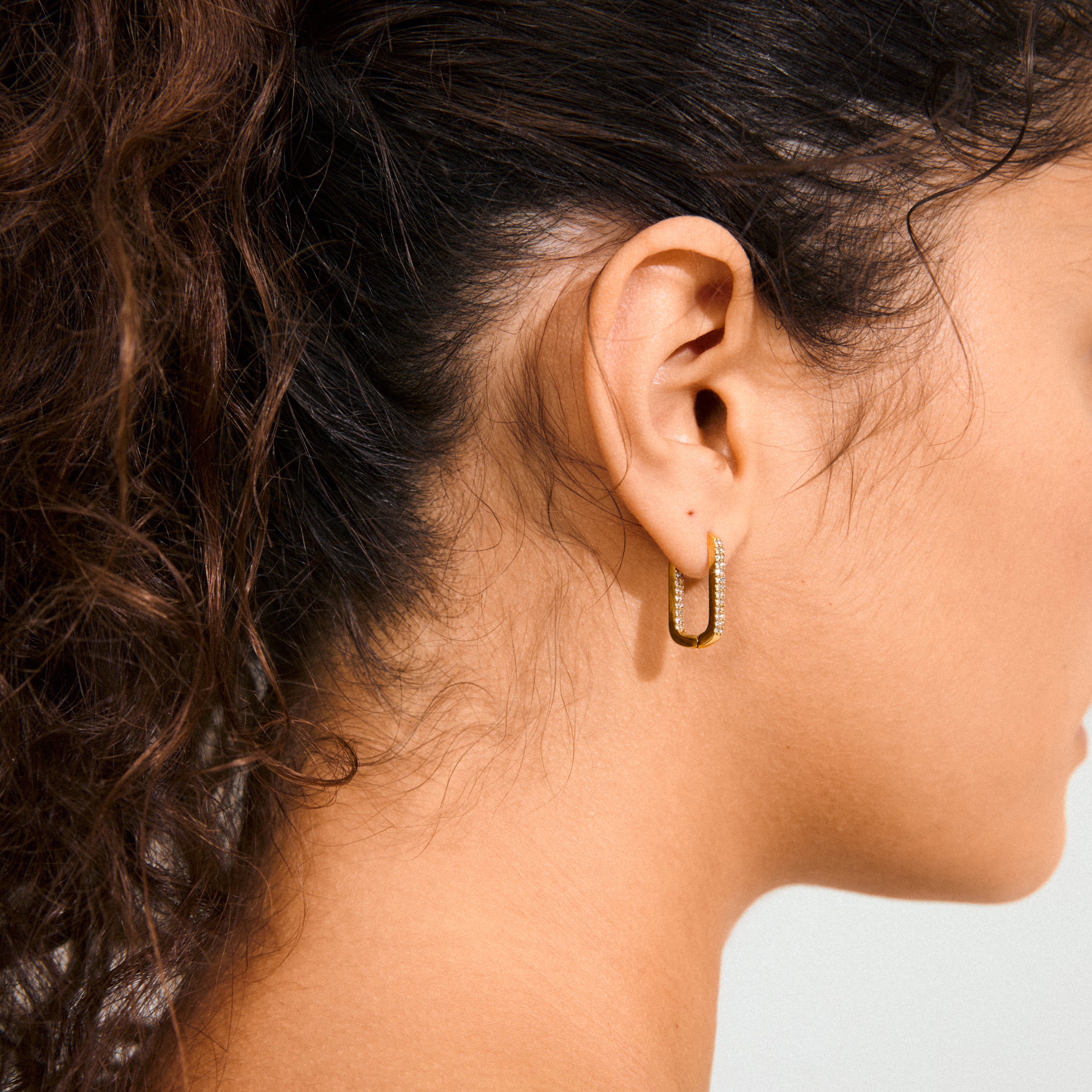 Close-up of a gold-plated PHASE oval hoop earring by Pilgrim, featuring sparkling Preciosa crystals, worn on a person's ear with hair pulled back.
