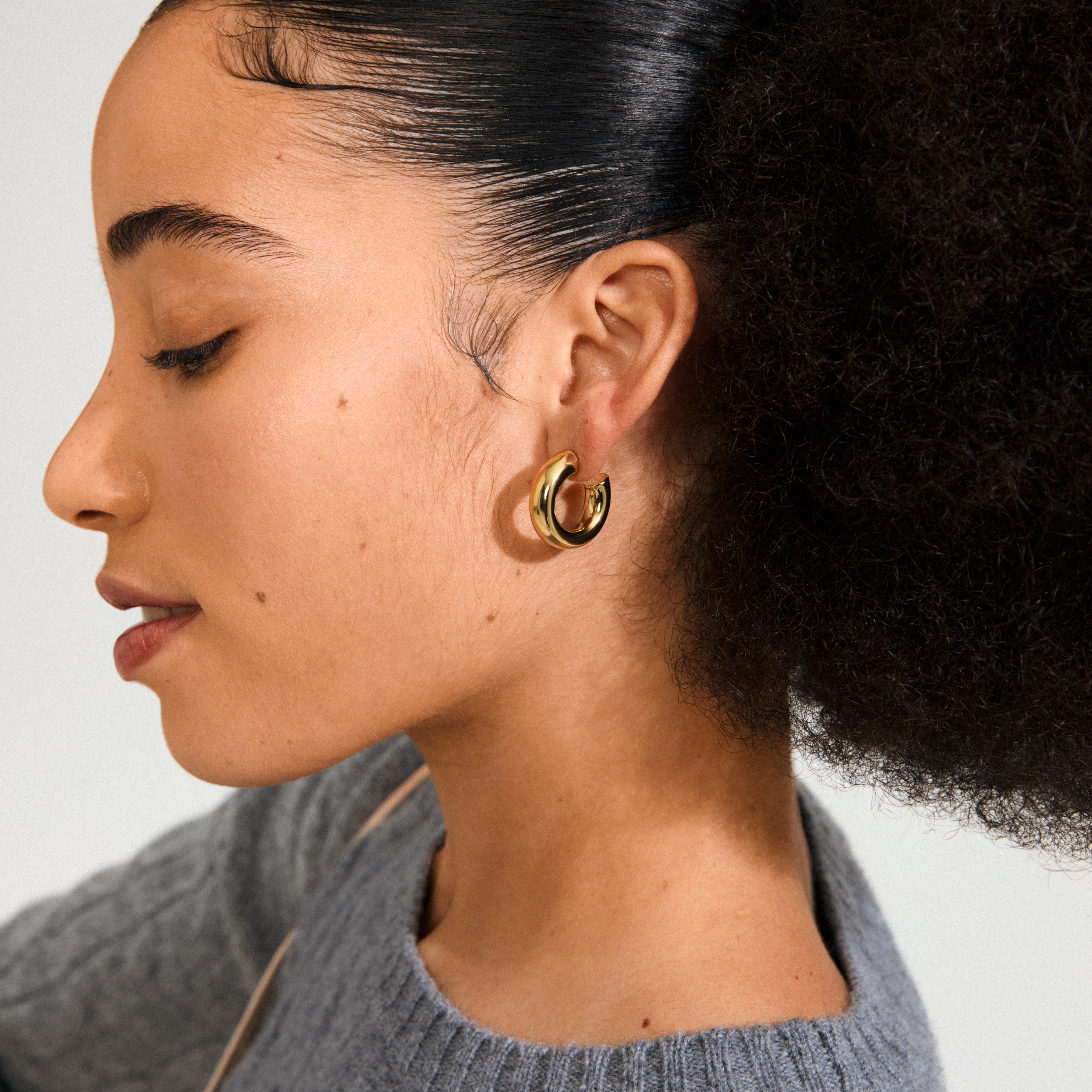 Side profile showing a person wearing PHASE large hoops, gold-plated, with textured hair and gray knit top, embodying Pilgrim's blend of classic and trendy style.