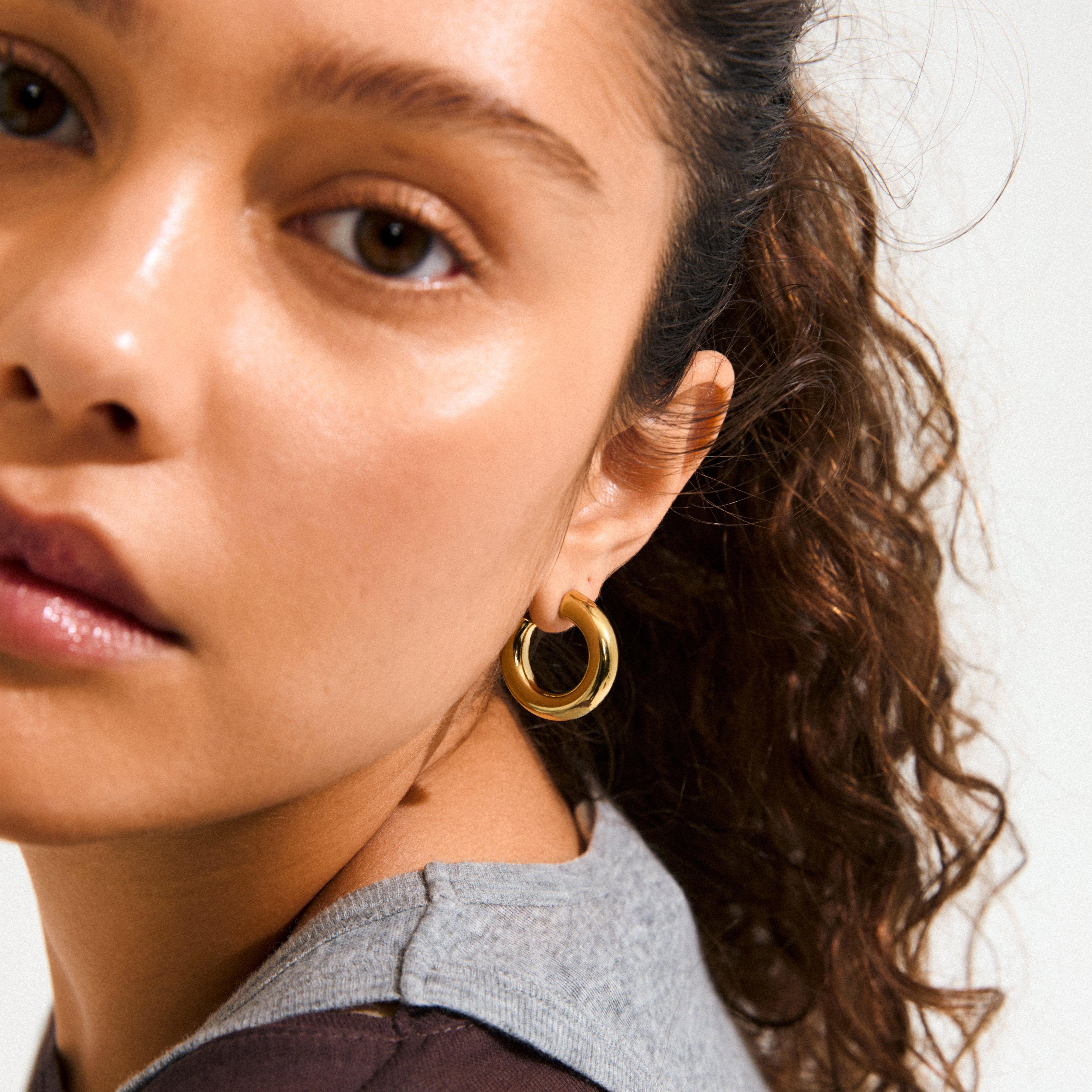 Young woman wearing PHASE medium gold-plated hoops by Pilgrim, showcasing a chunky silhouette. The earring complements her layered outfit, embodying quiet luxury and autumn style.