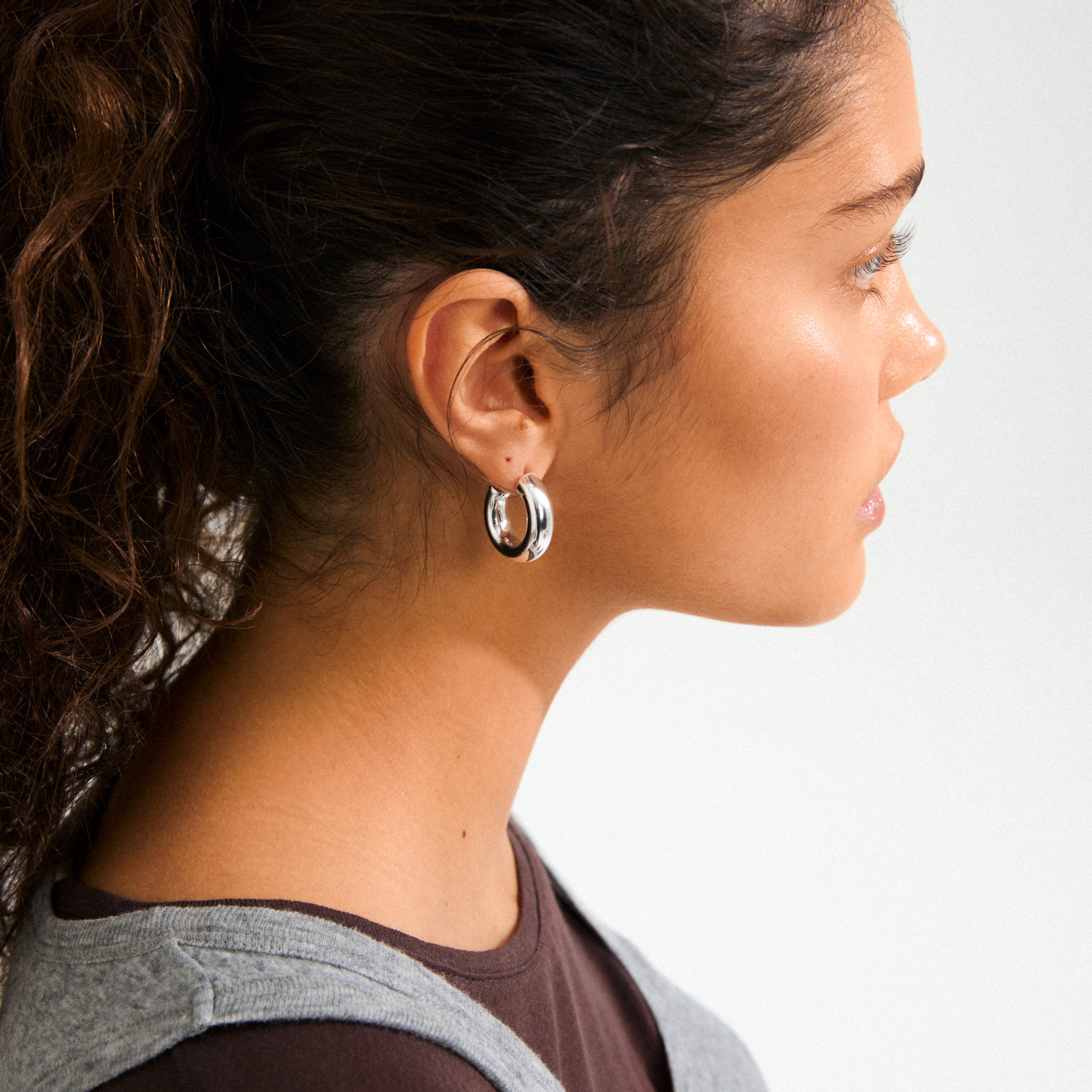 Side-profile of a young woman wearing Pilgrim's PHASE medium silver-plated hoops, showcasing their chunky, elegant design complementing her sleek, minimalist attire.