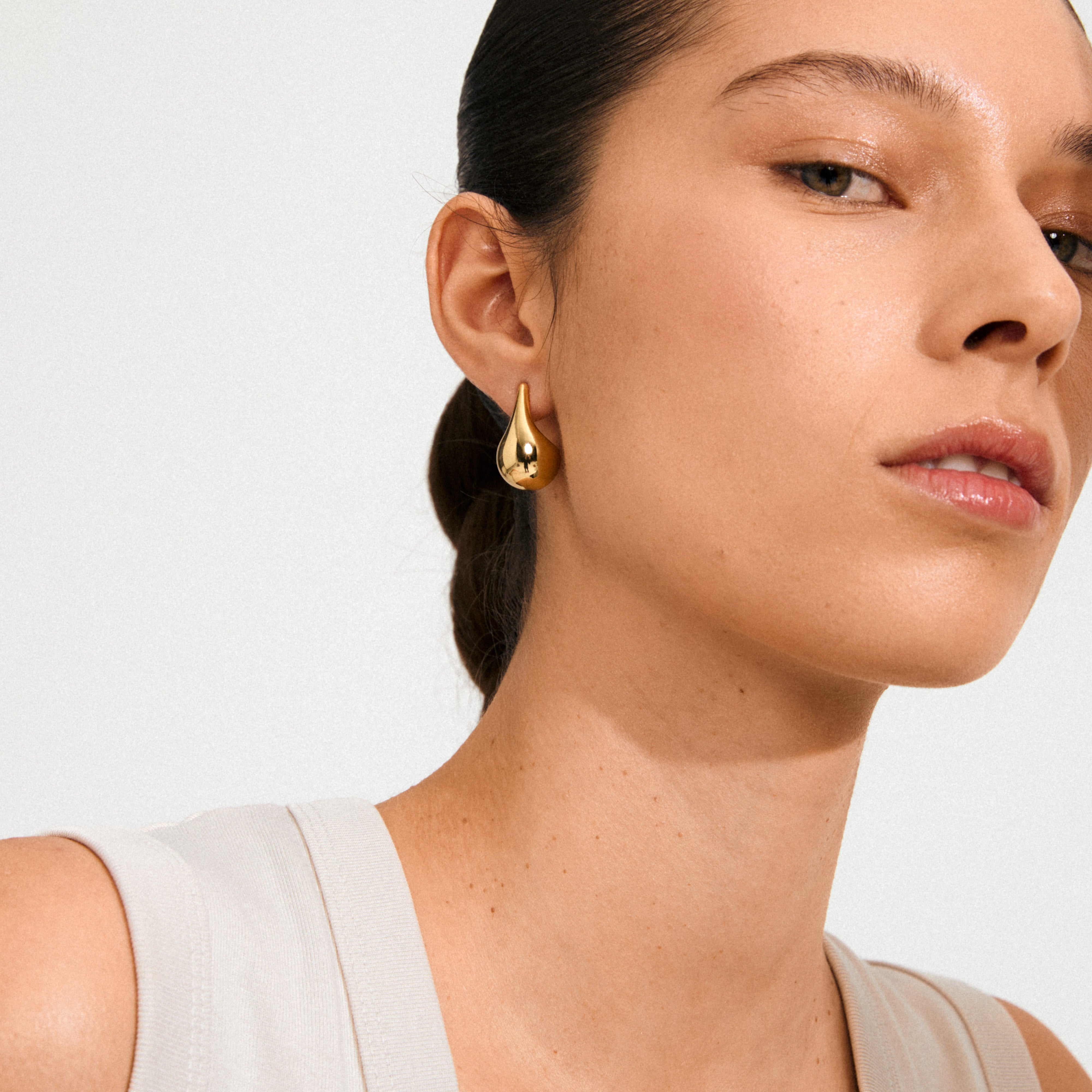 Woman wearing JUNIPER earrings, gold-plated teardrop design by Pilgrim, showcasing minimalist elegance and Scandinavian style against a softly lit background.