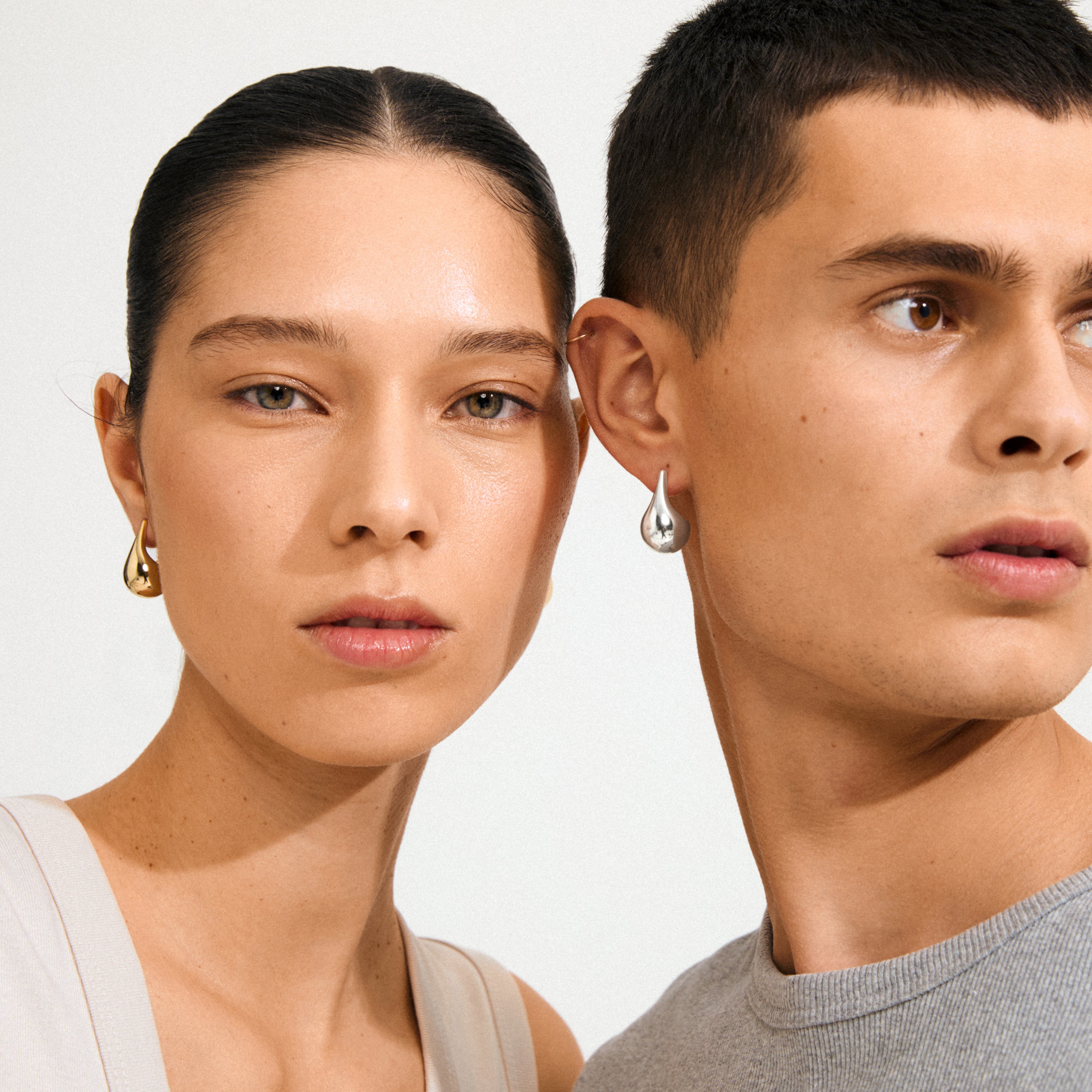 Two young adults showcase Pilgrim's JUNIPER earrings: one gold-plated and one silver, both teardrop-shaped, embodying Scandinavian elegance and modern style.