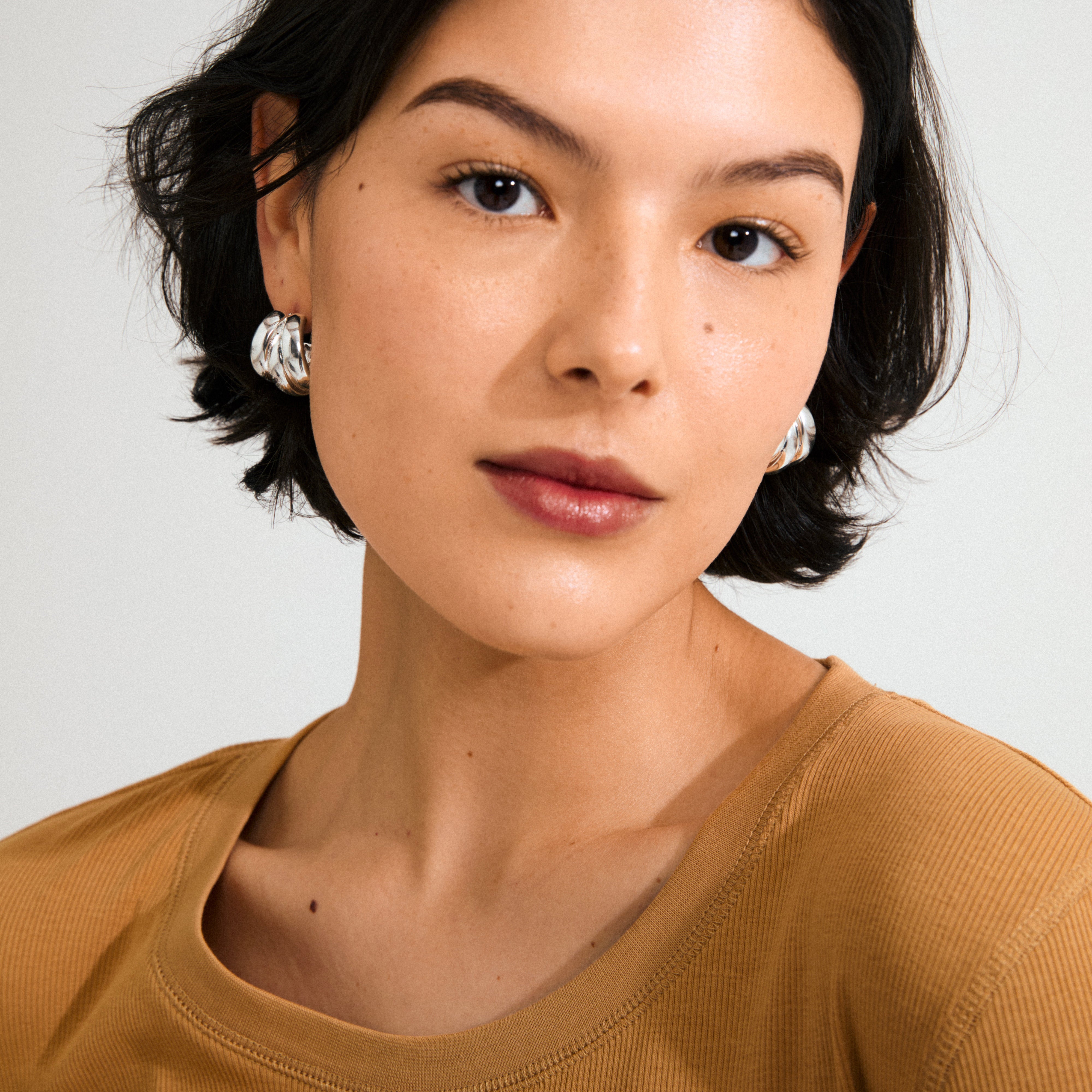 Young person wearing Pilgrim's silver-plated DAMIAN earrings, featuring a chunky triple-ring design. Close-up emphasizes the earrings' bold, trendy style against minimalist makeup.