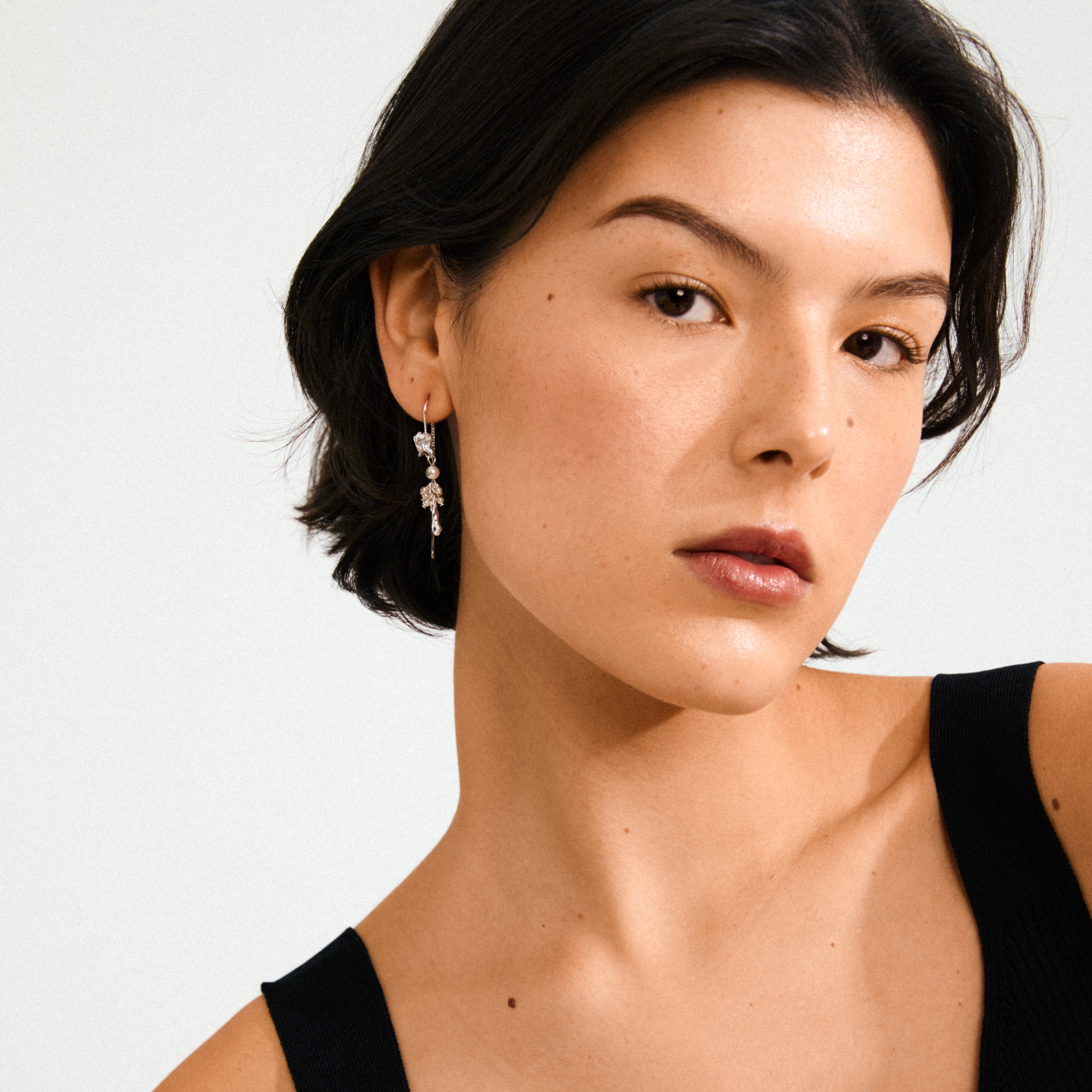 Young woman wearing MILO chain earrings; silver-plated, featuring mother-of-pearl beads, resembling delicate water bubbles, adding a playful, adventurous touch against a simple black-strap top.