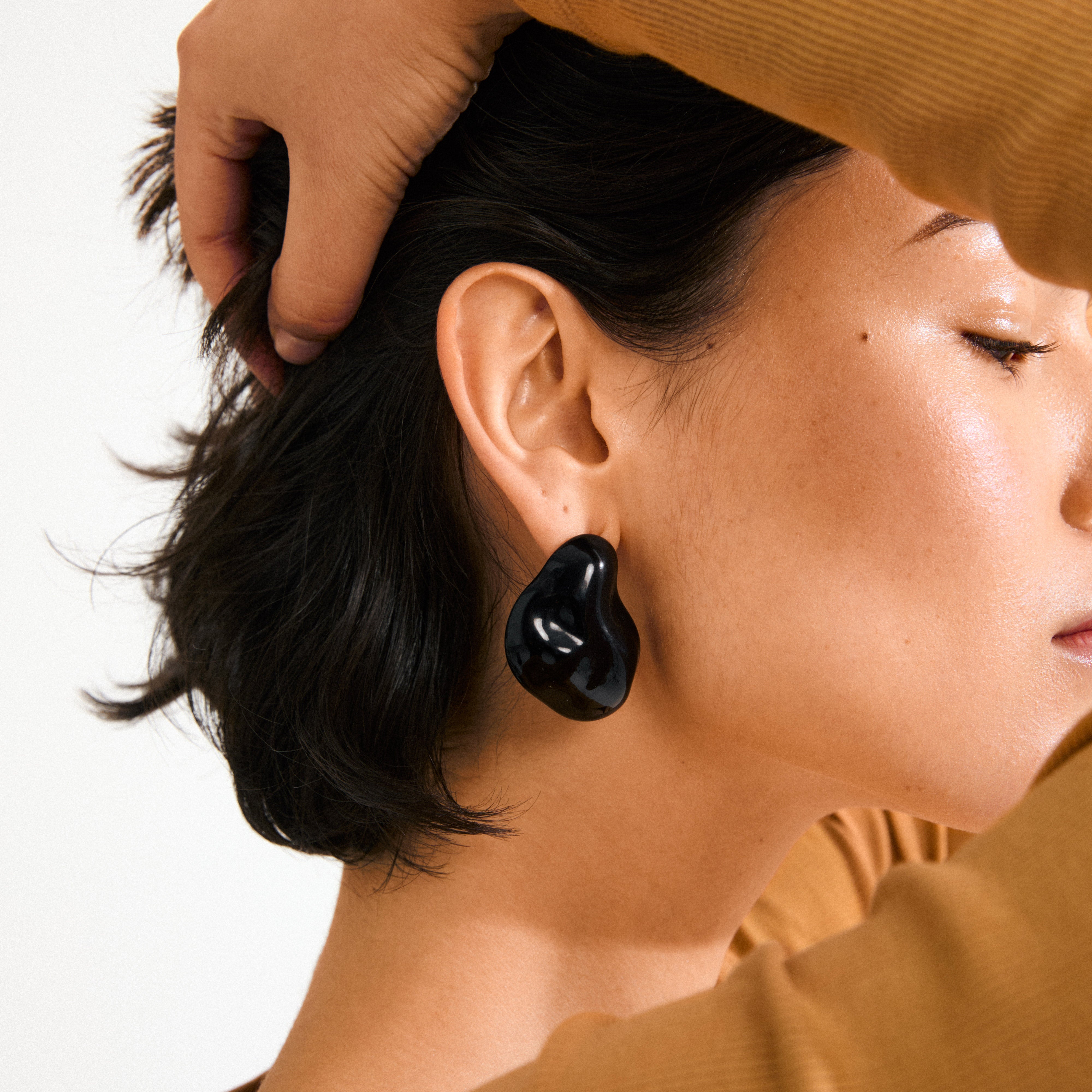 Side view of a person wearing Pilgrim's LIAN earrings in glossy black, showcasing their bold, pebble-like design against smooth skin and a camel top.