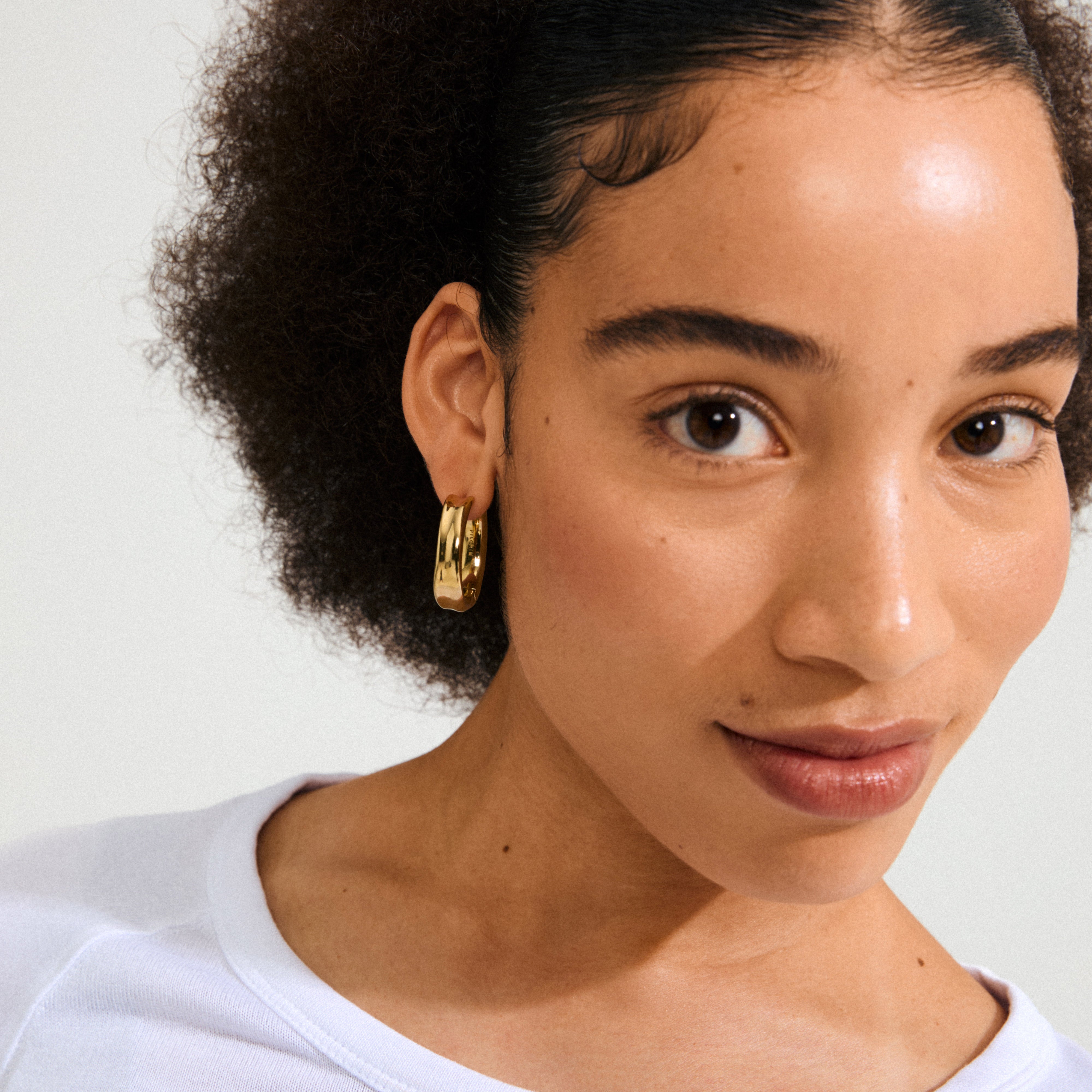Young woman wearing Pilgrim's KAYDEN gold-plated hoops, showcasing their broad, shiny design. Her natural afro and simple white top highlight the earrings' versatility.