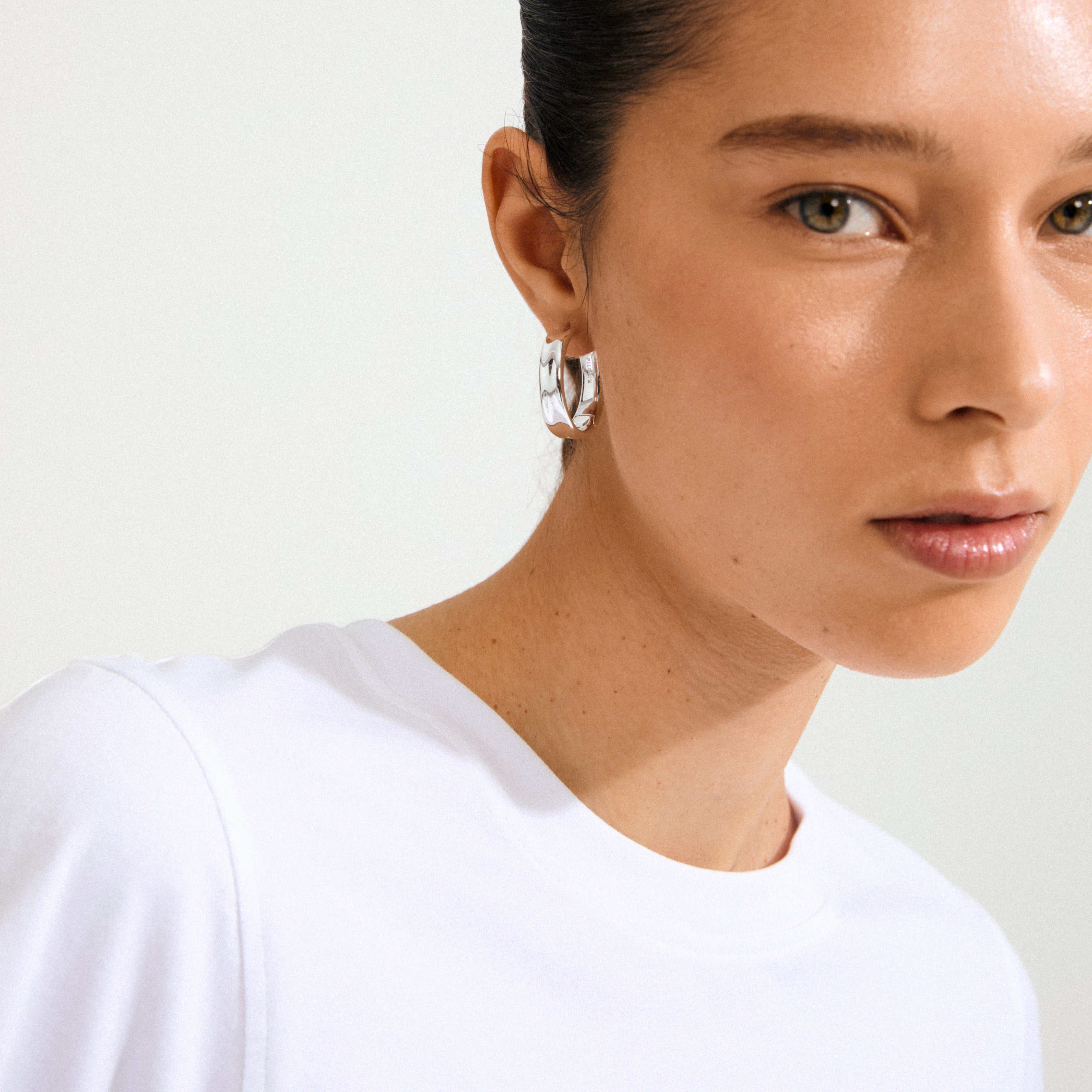 Young woman wearing a KAYDEN hoops silver-plated earring, showcasing its broad, polished design against a simple white T-shirt. Hair pulled back, highlighting elegance.