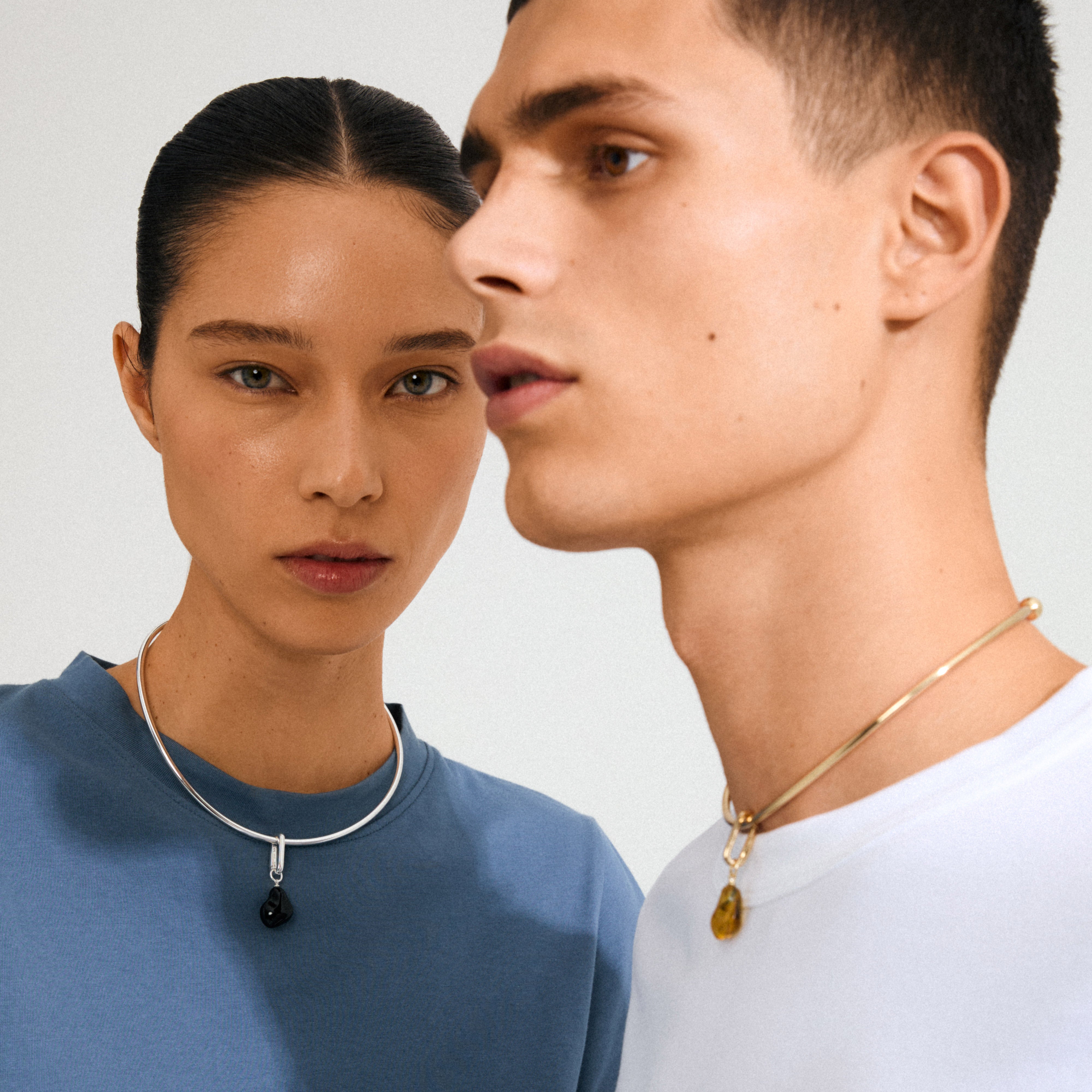 Models showcasing Pilgrim's CHARM mold pendant necklaces: a silver-plated black stone on her, and a gold-banded amber-tone stone on him. Modern, minimalist style.